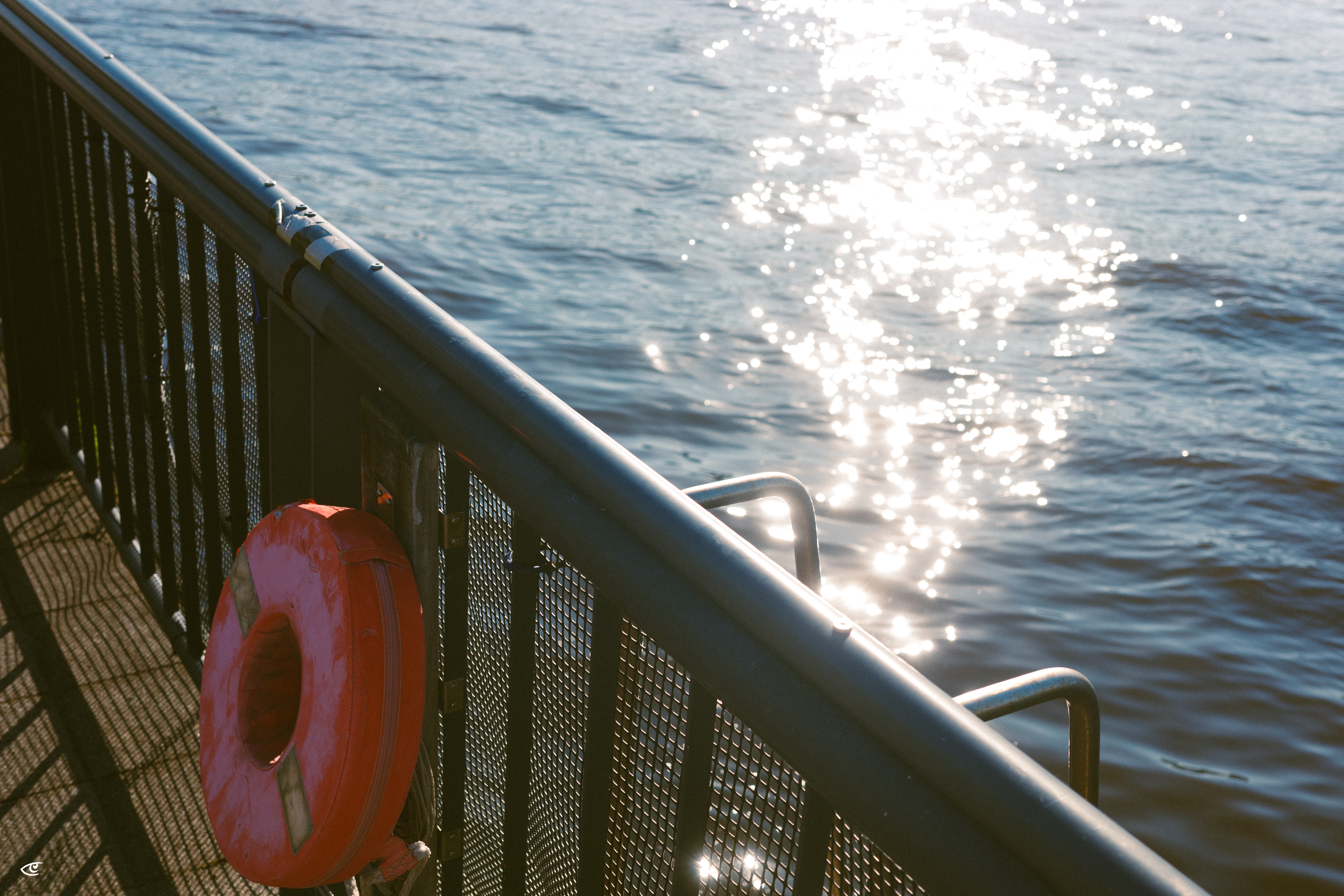 Red life ring attached to a black metal railing on a waterfront walkway, with sunlight reflecting off rippling water in the background.