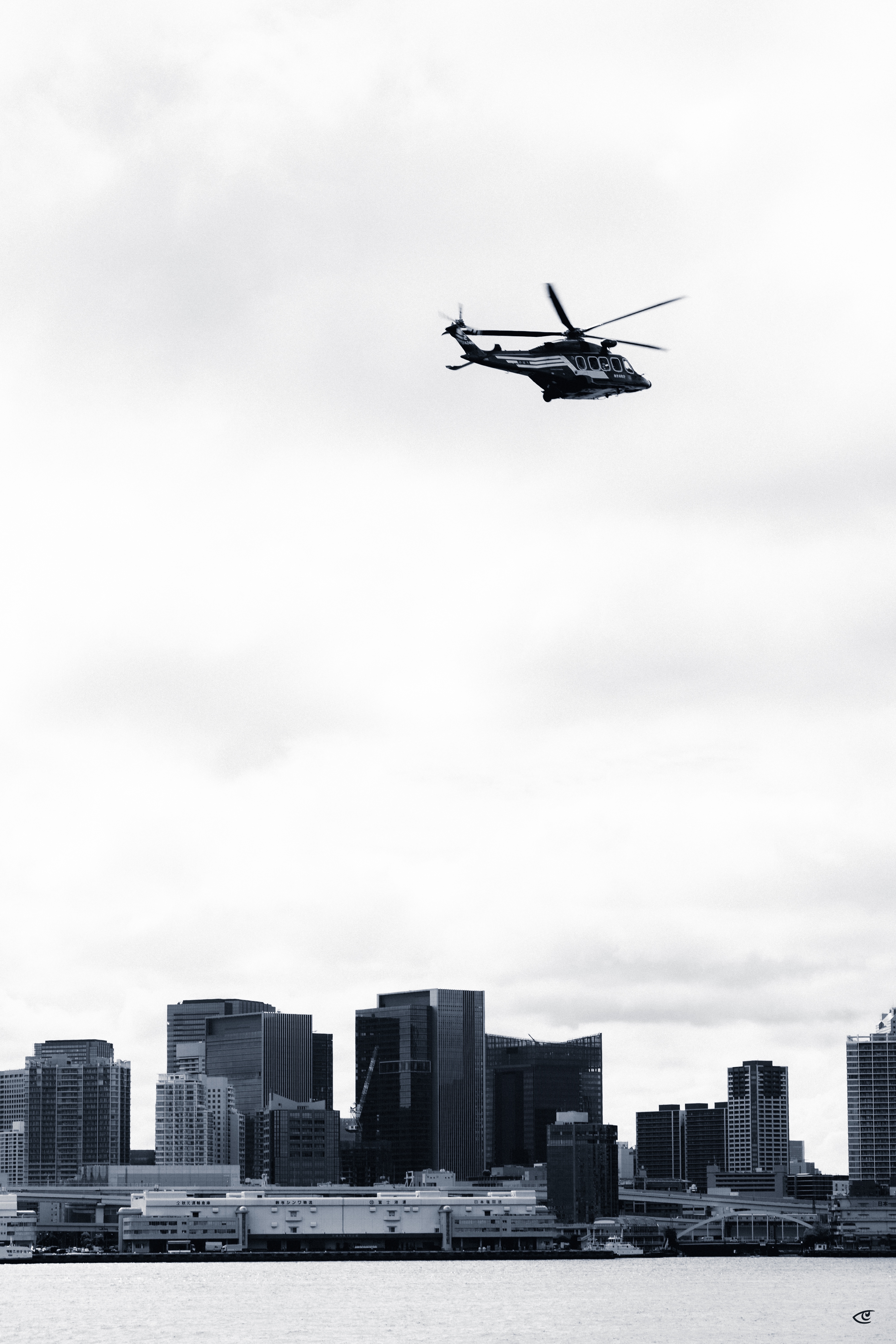 Black helicopter flying above a city skyline, with high-rise buildings along a waterfront under an overcast sky.
