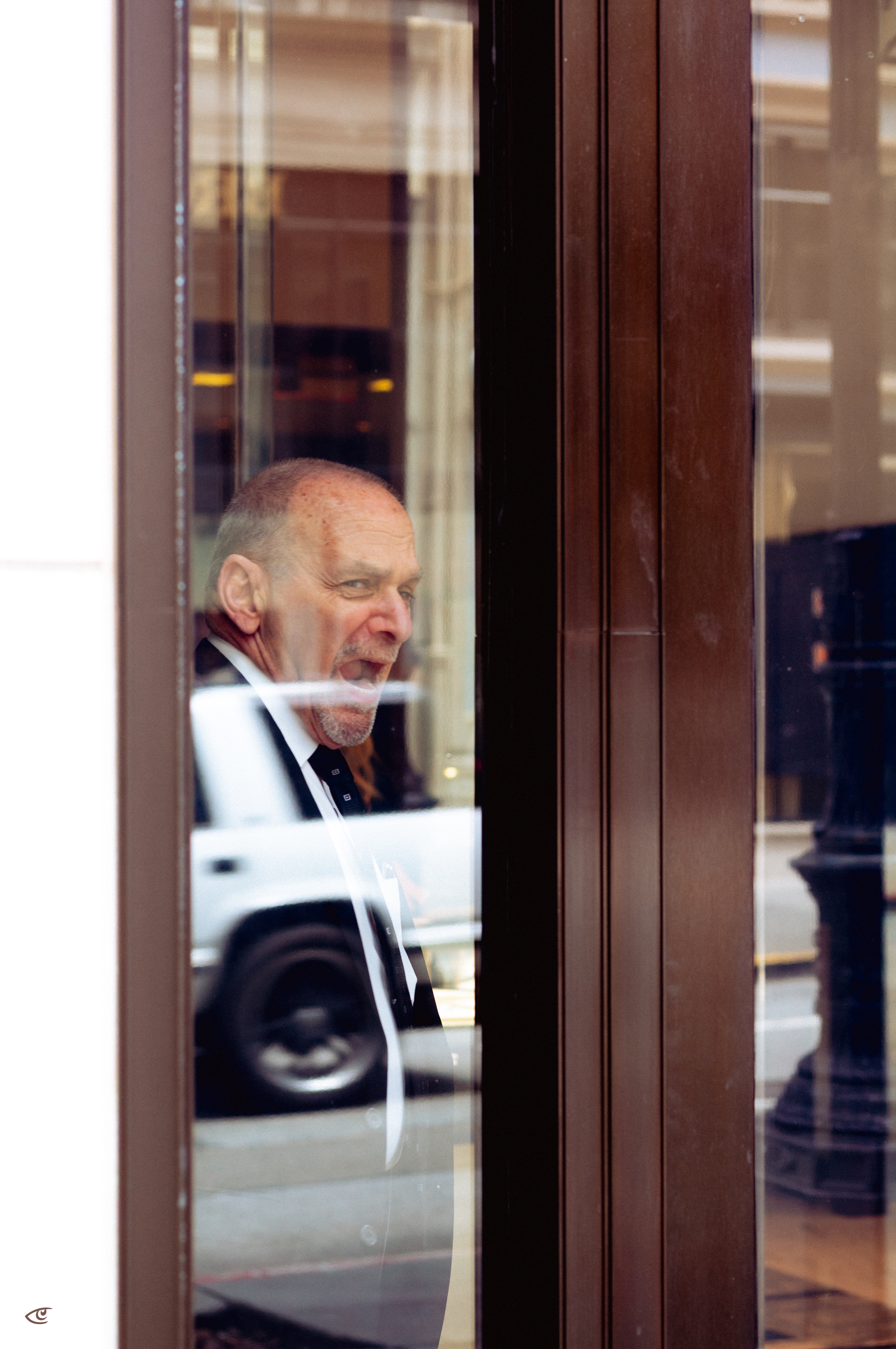 Older man in a dark suit and tie seen through a glass storefront door, with street reflections including a passing vehicle and a lamppost across the glass.