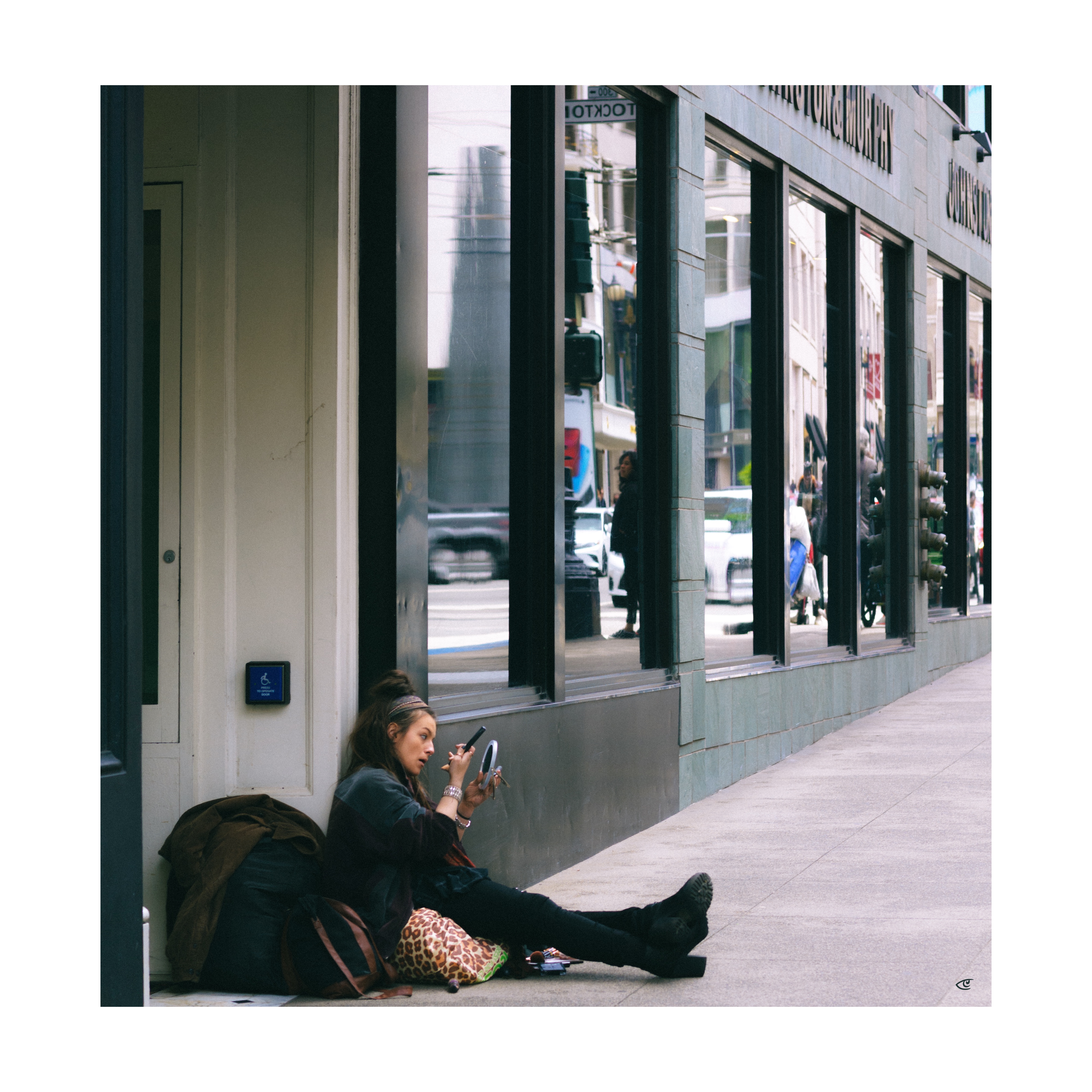 Person sitting on a city sidewalk against a building wall, holding a small round mirror and applying makeup; bags and belongings beside them, with large storefront windows reflecting street traffic and pedestrians.