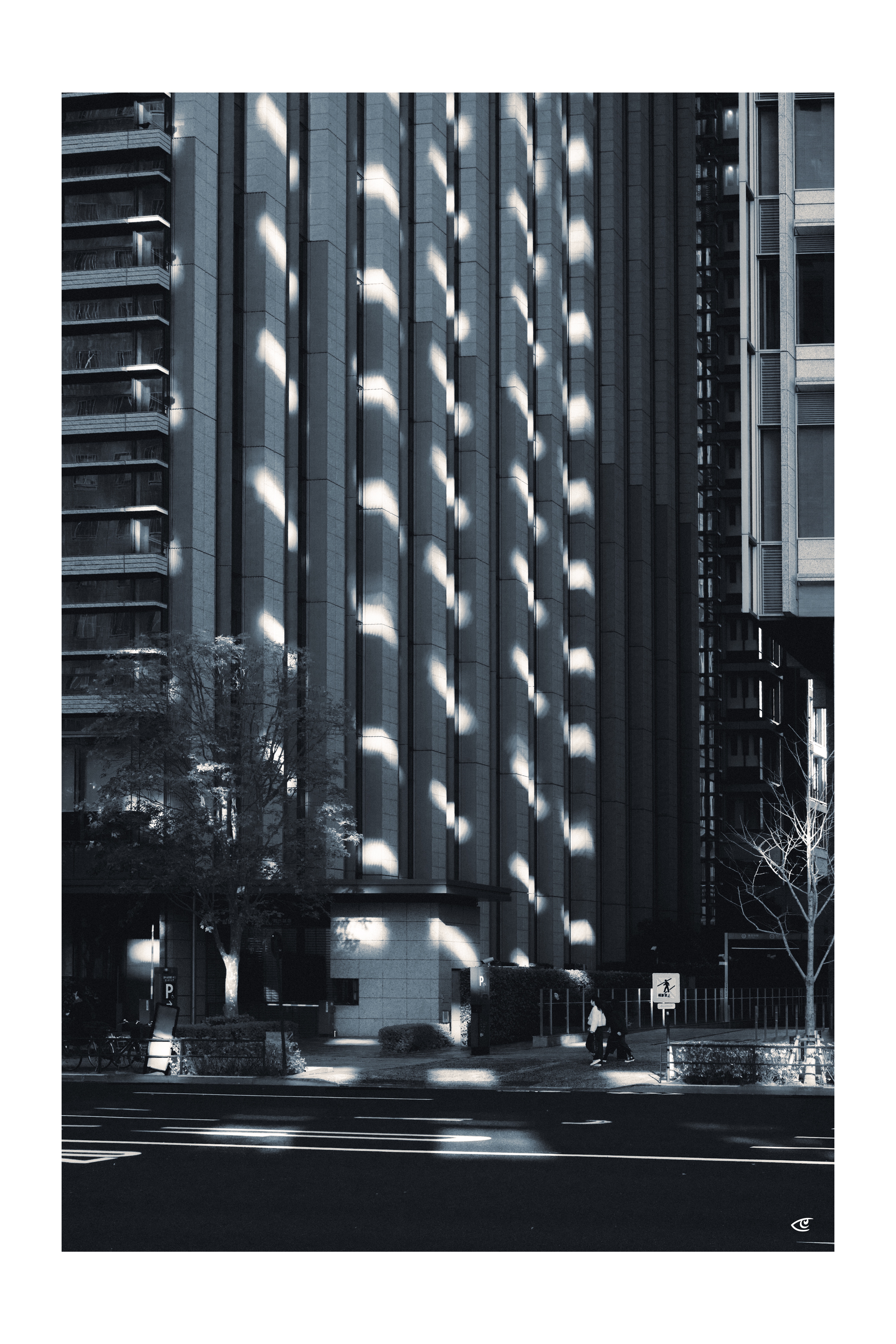 Tall urban building facade with vertical concrete columns and scattered sunlit patches; apartment balconies on the left, a narrow tower section on the right, and a street-level entrance with trees, a crosswalk sign, and a few pedestrians at the bottom.