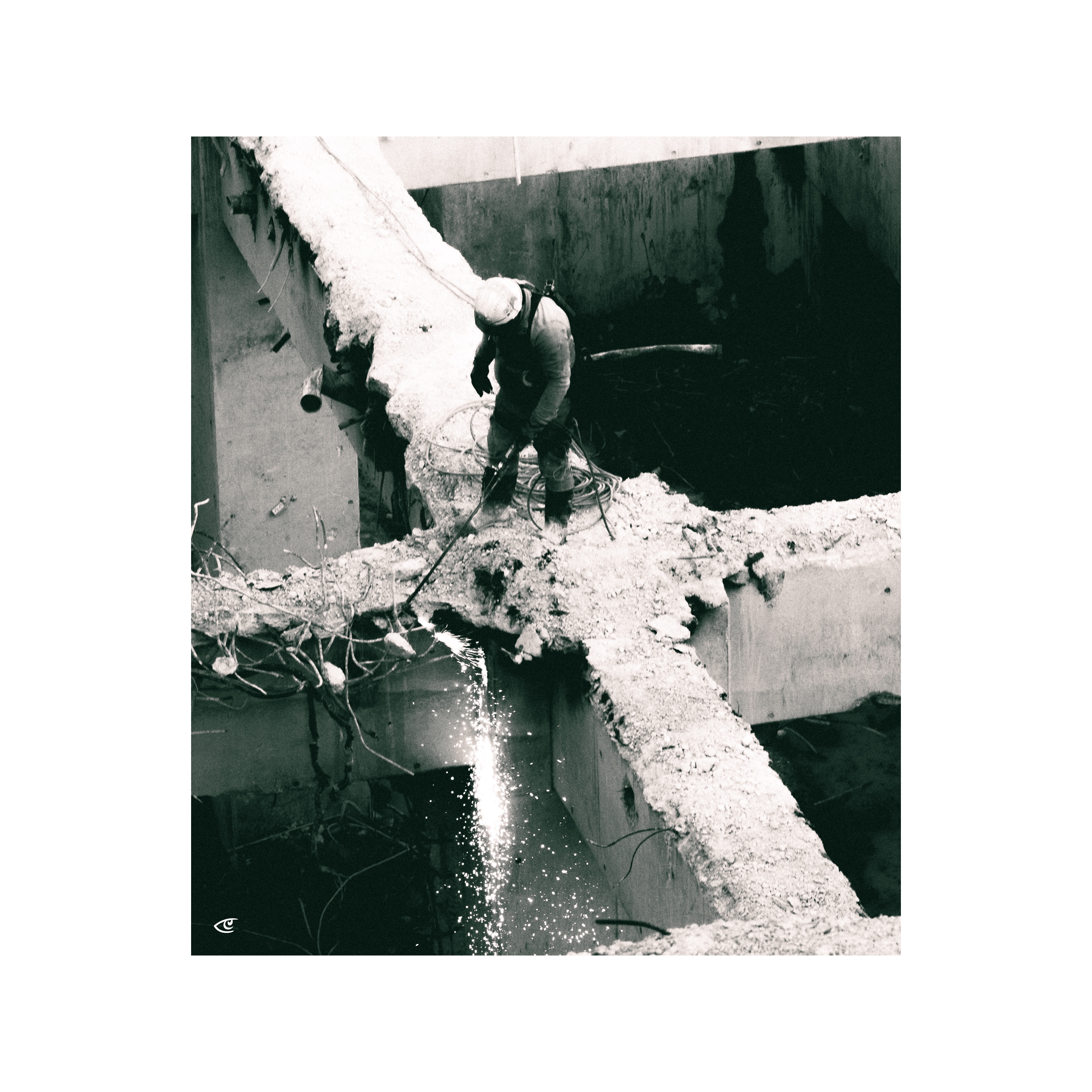 Worker wearing a hard hat stands on a broken concrete beam, using a cutting tool that throws sparks downward into a large, dark opening; exposed rebar and rubble surround the partially demolished structure.