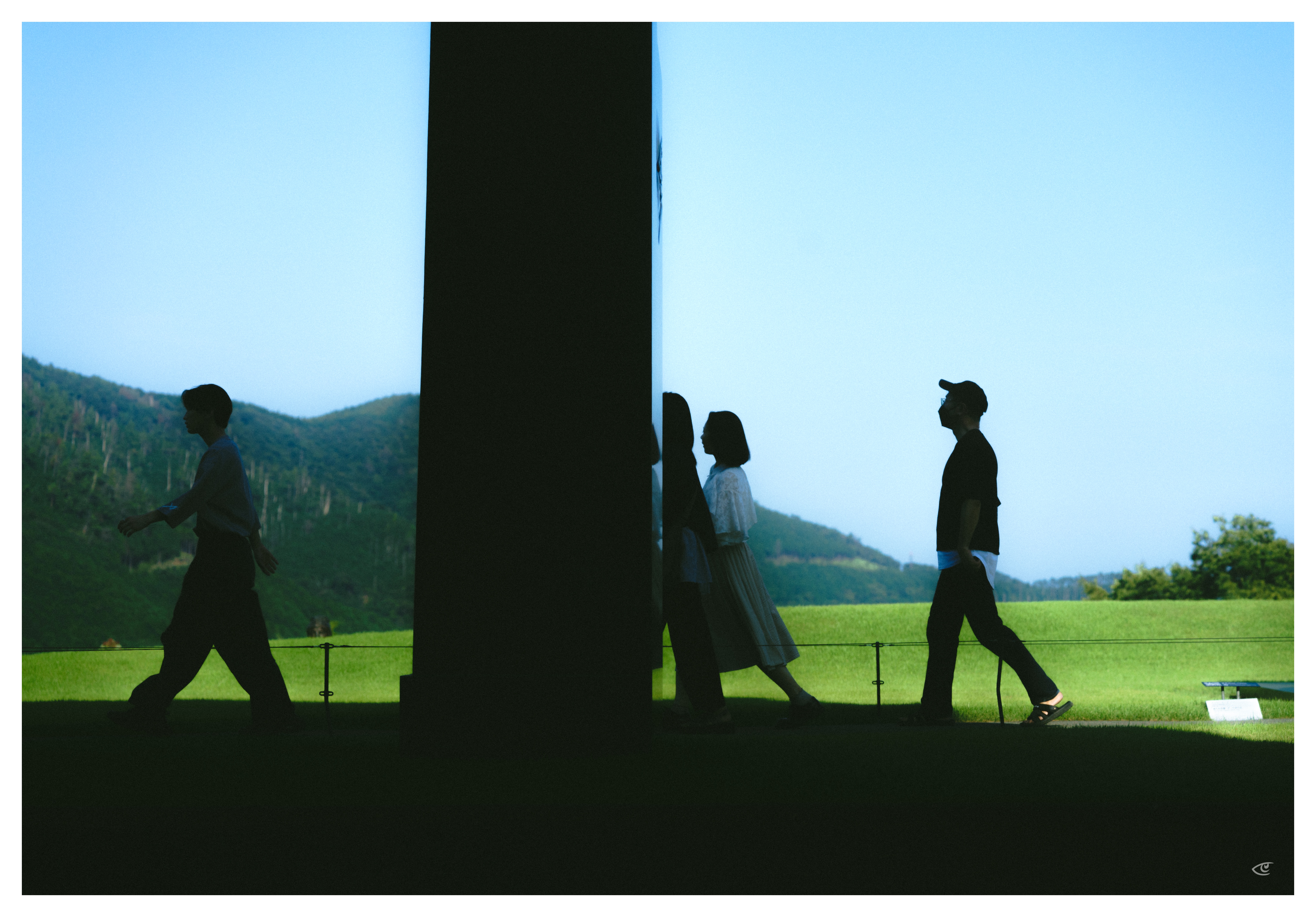 Silhouettes of four people walking in opposite directions beneath a large dark structure, with a green field and tree-covered mountains in the background under a clear sky.