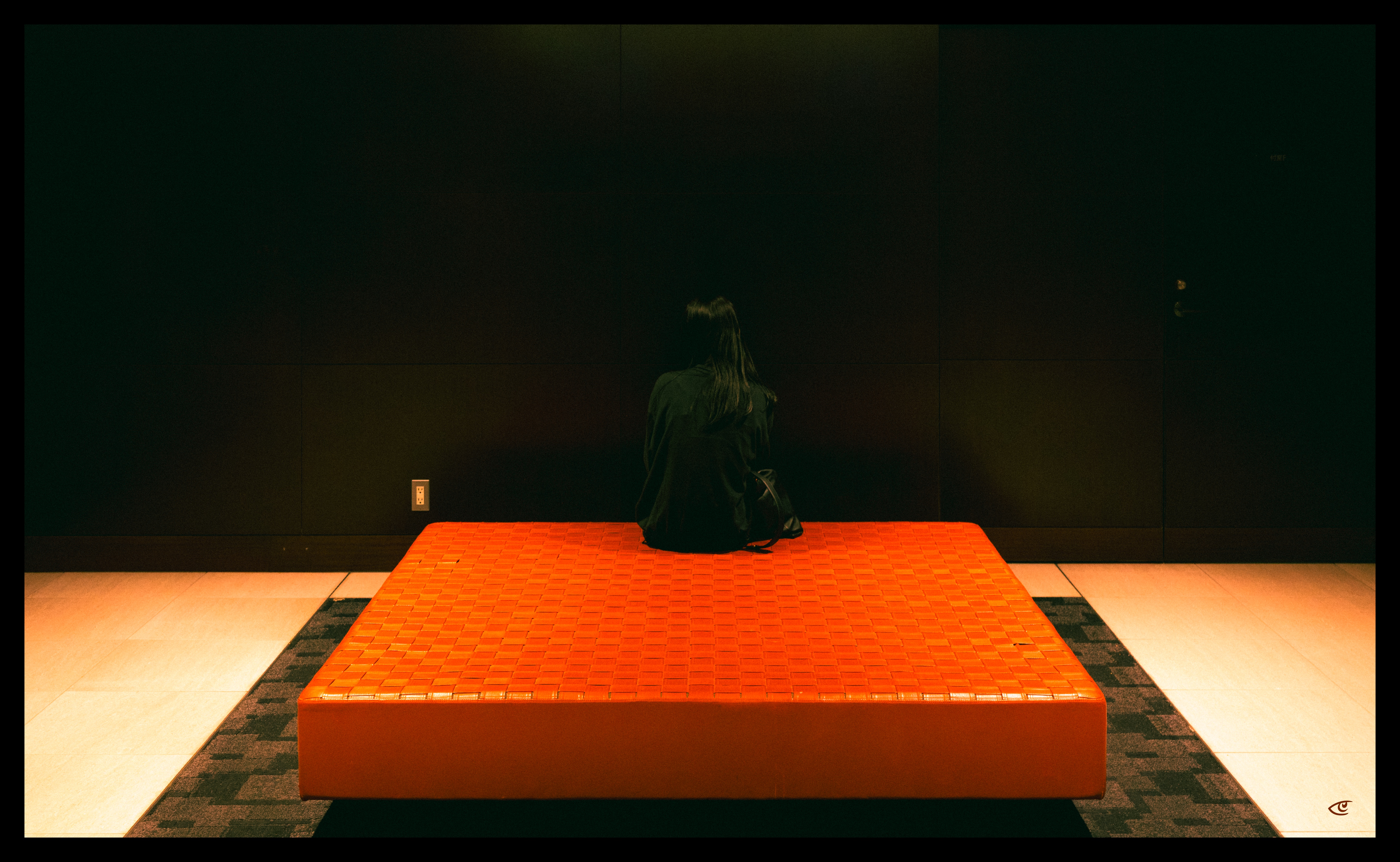 Person in dark clothing sitting with their back to the camera on a low, bright orange square platform centered in a dim room with a black wall and patterned carpet border.