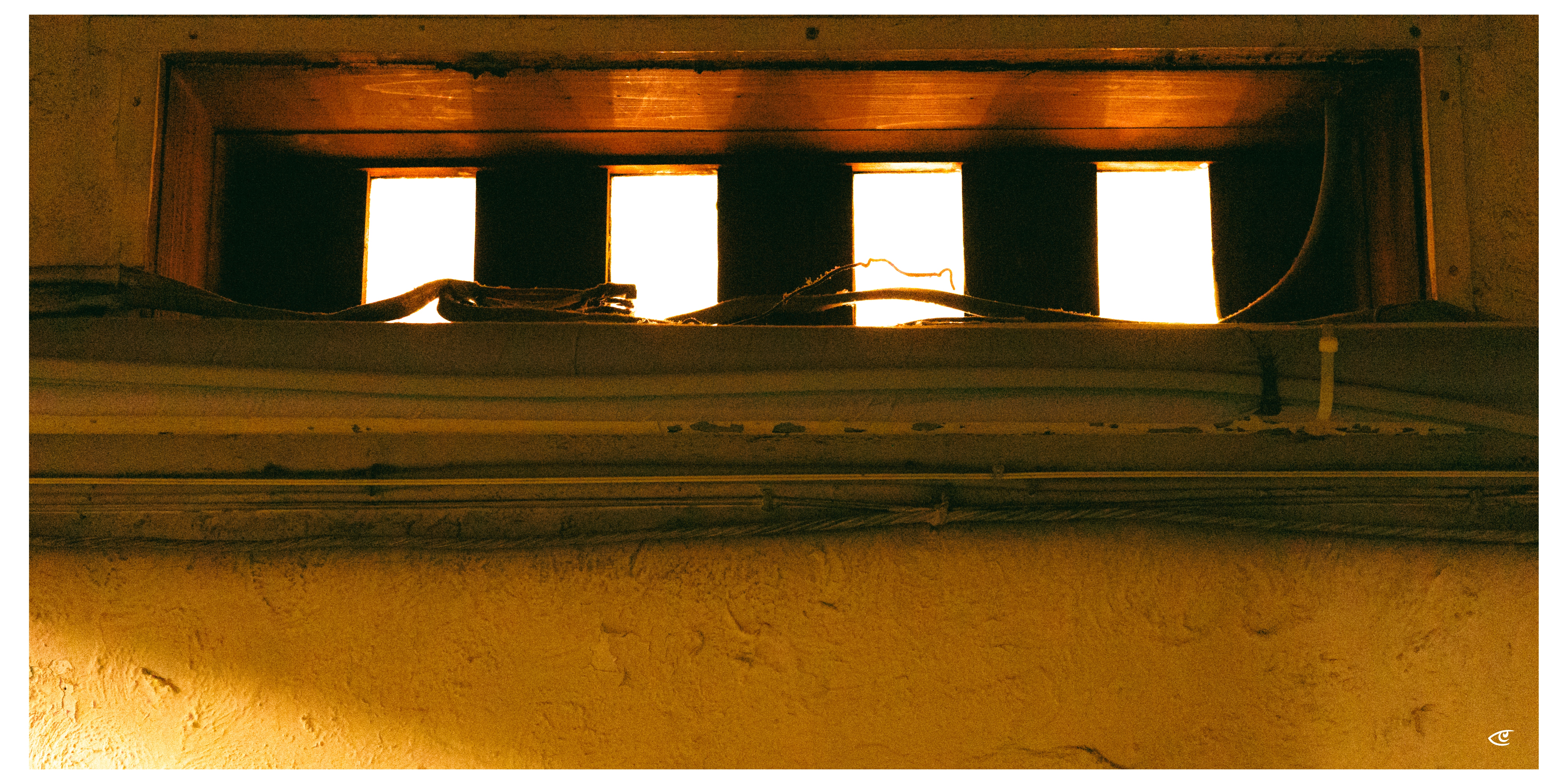 Four narrow vertical window openings near the top of a textured beige wall, with bright light shining through and cables resting along the sill inside a recessed frame.