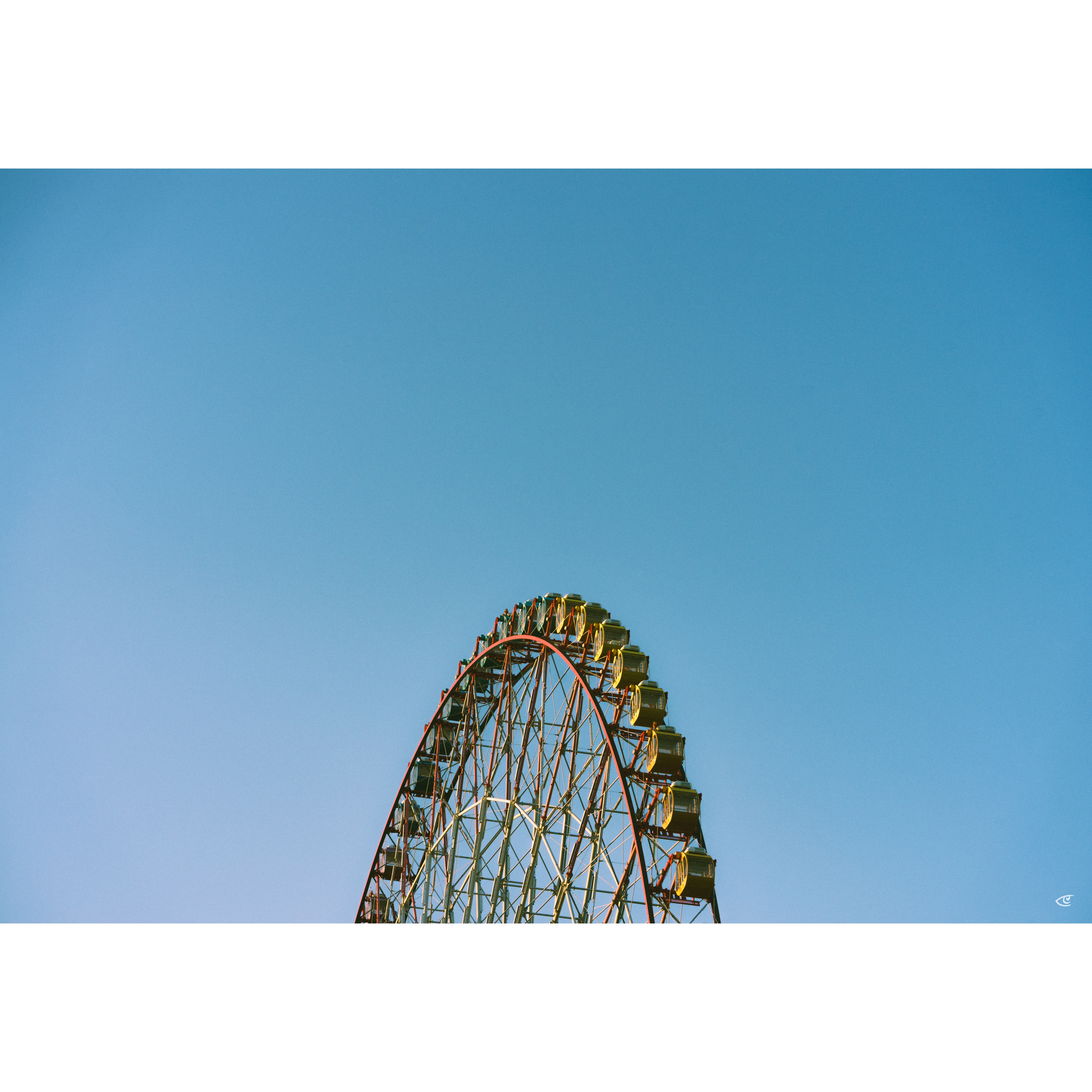 Upper portion of a Ferris wheel with yellow gondolas rises from the bottom edge against a clear blue sky, with large empty sky space above.