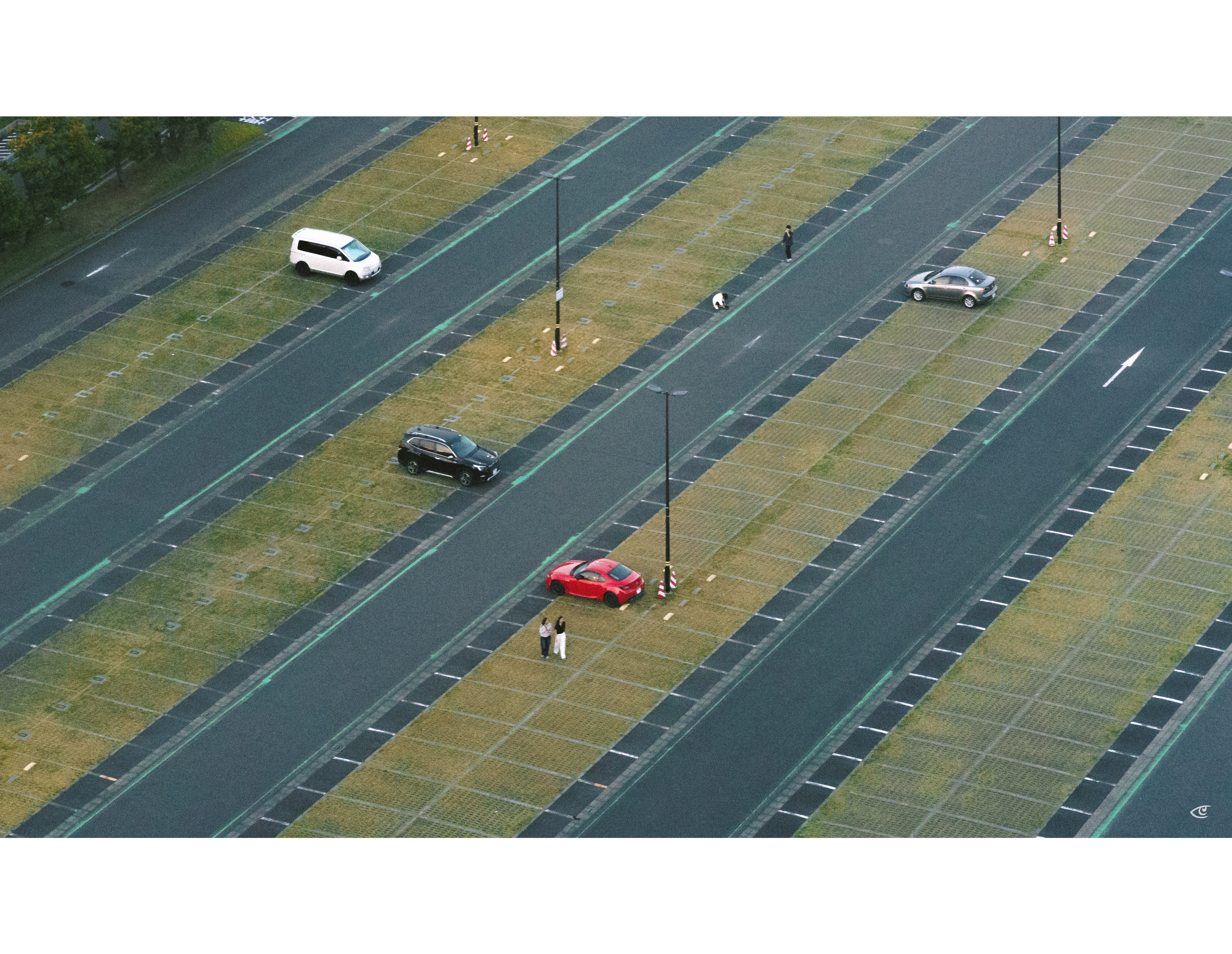Aerial view of a wide multi-lane roadway with grassy median strips and tall light poles, showing a white van, black car, red car, and gray car stopped in separate lanes, with a few pedestrians standing and walking between lanes.