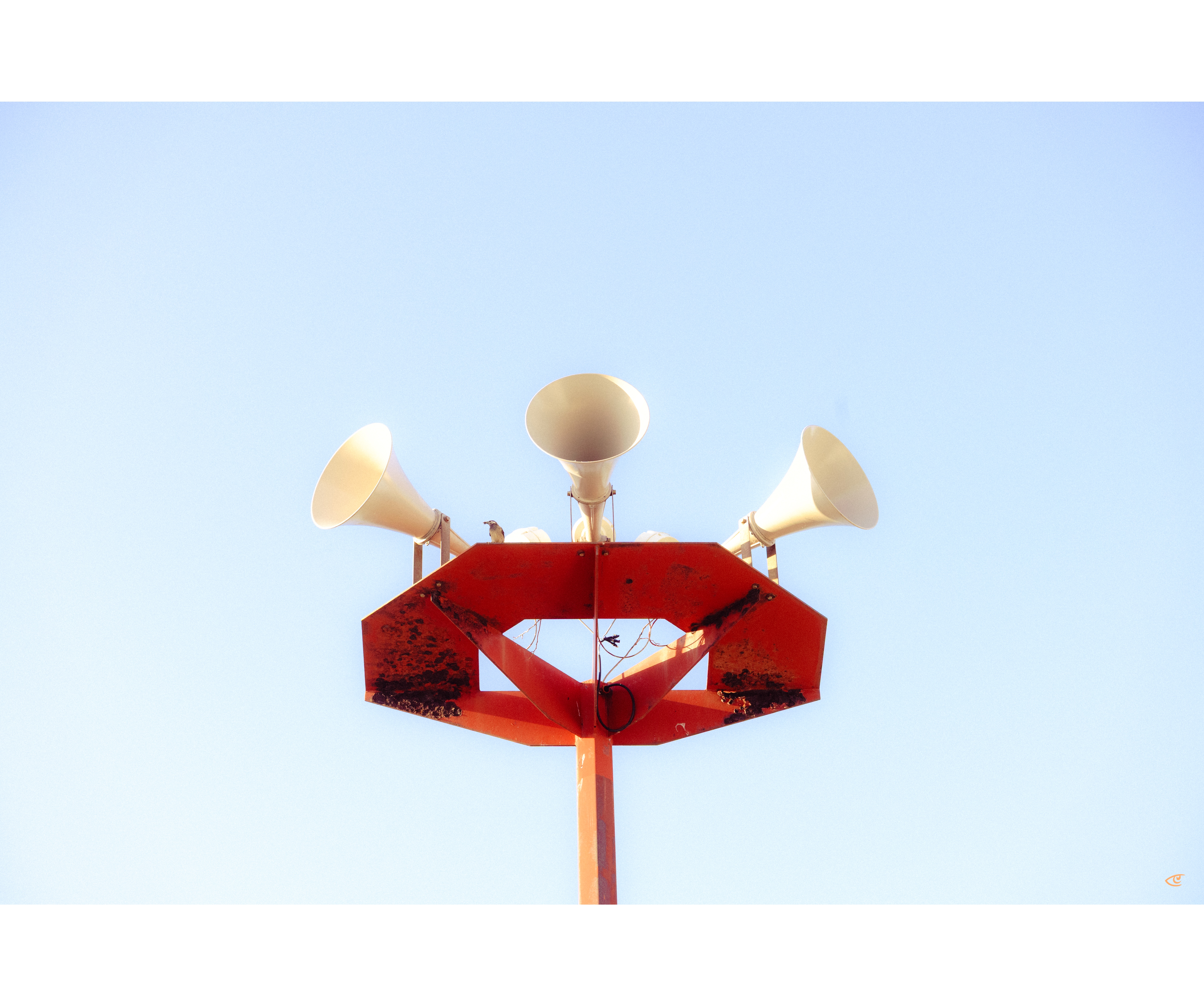 Three white horn loudspeakers mounted on a red metal platform atop a tall pole, viewed from below against a clear blue sky.