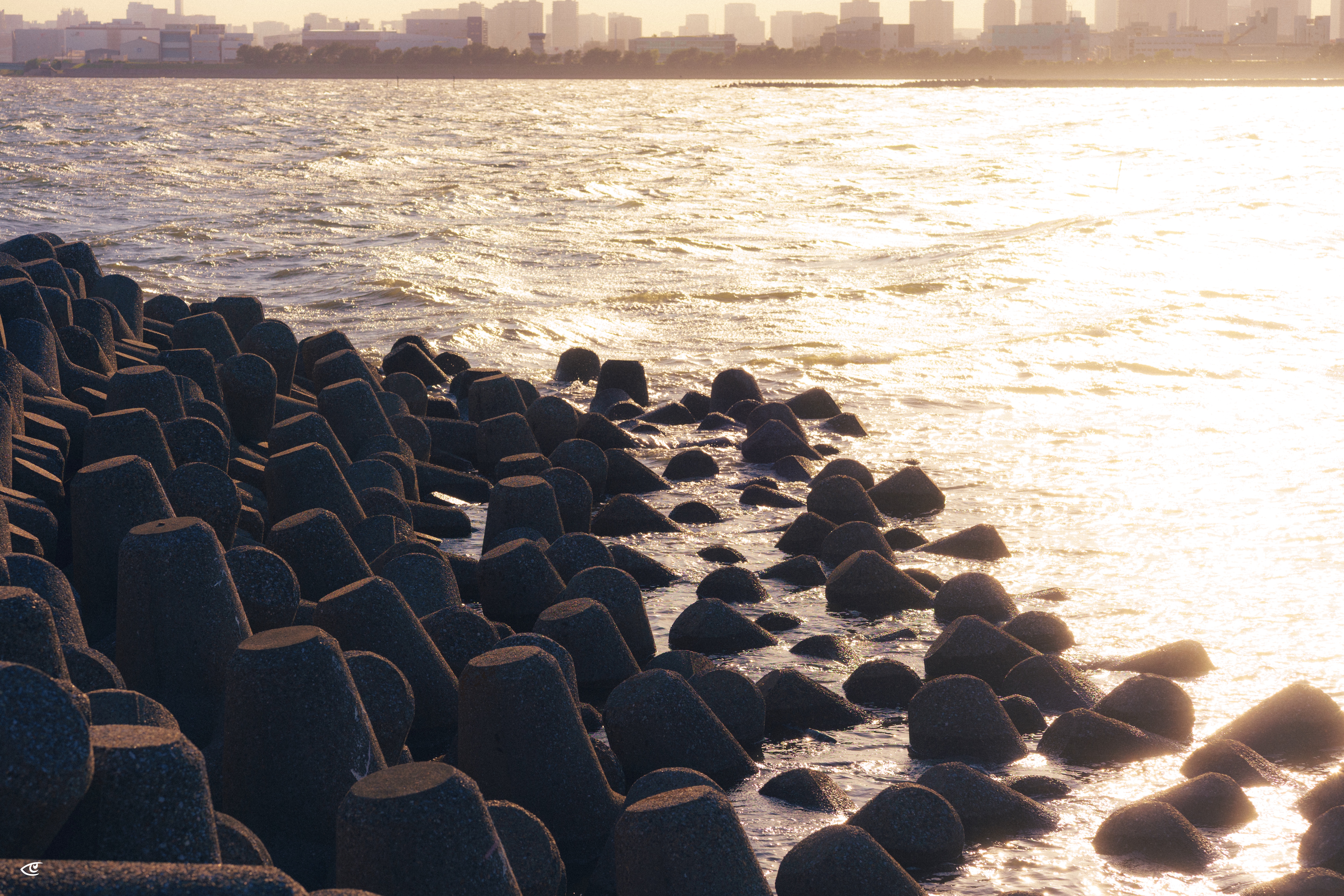 Concrete wave-breaker blocks along a shoreline with small waves washing between them, bright sun glare on the water to the right, and a distant city skyline on the horizon.