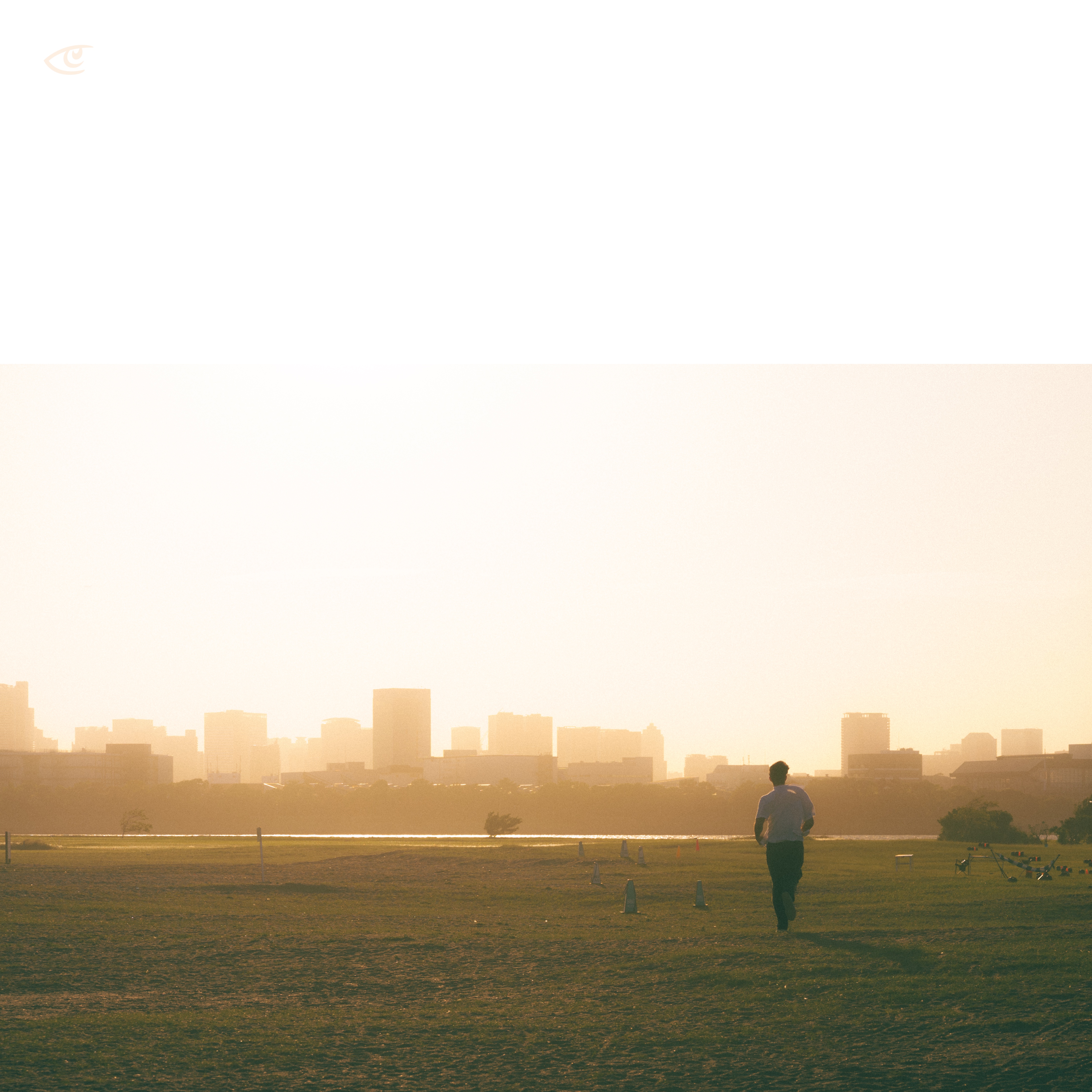 Person walking across an open grassy field at sunset, with long shadows, distant hazy city skyline on the horizon, and scattered cones and birds on the right side.