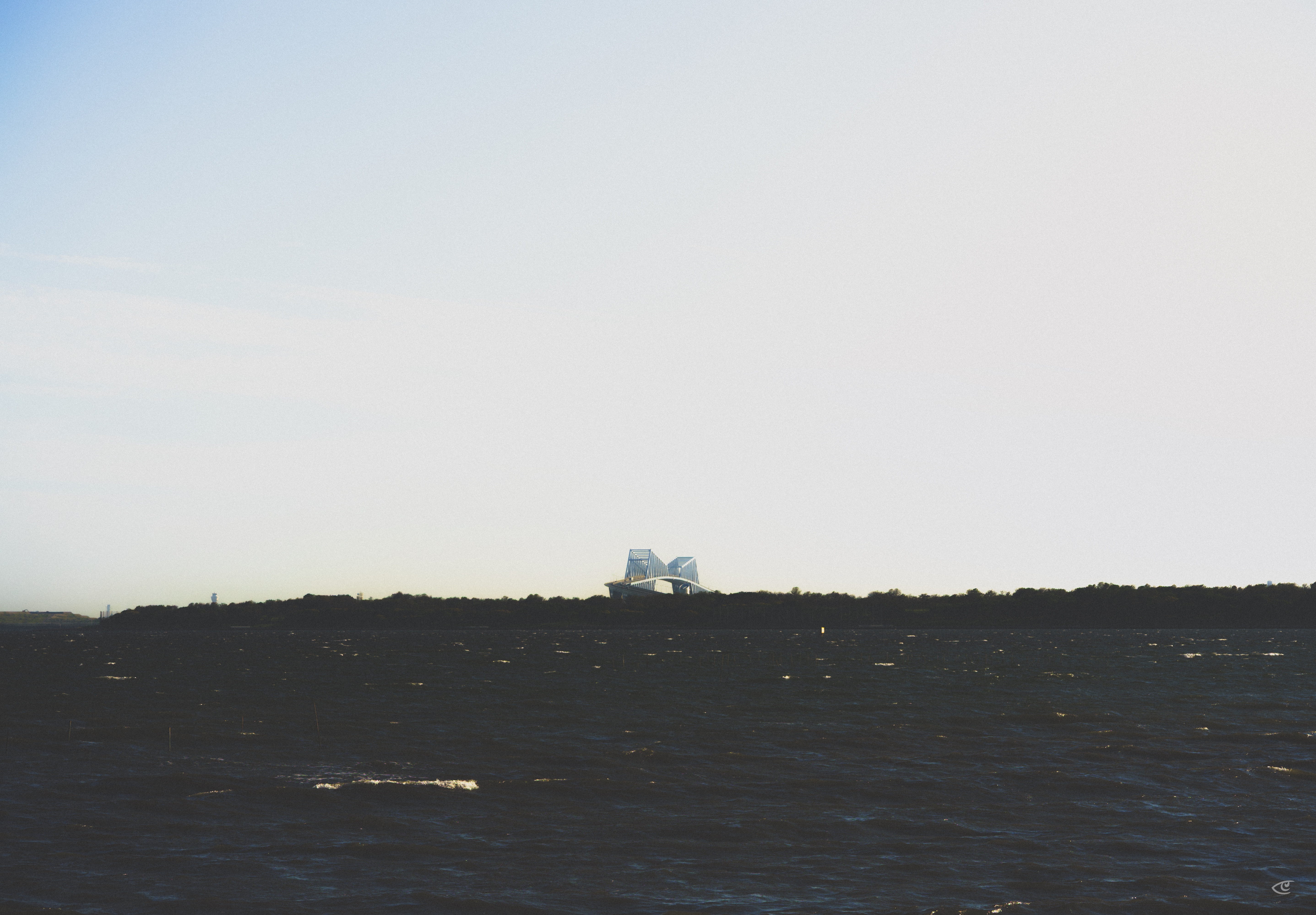 Distant white modern building on a low shoreline across dark, rippled water, with a pale blue sky filling most of the frame.