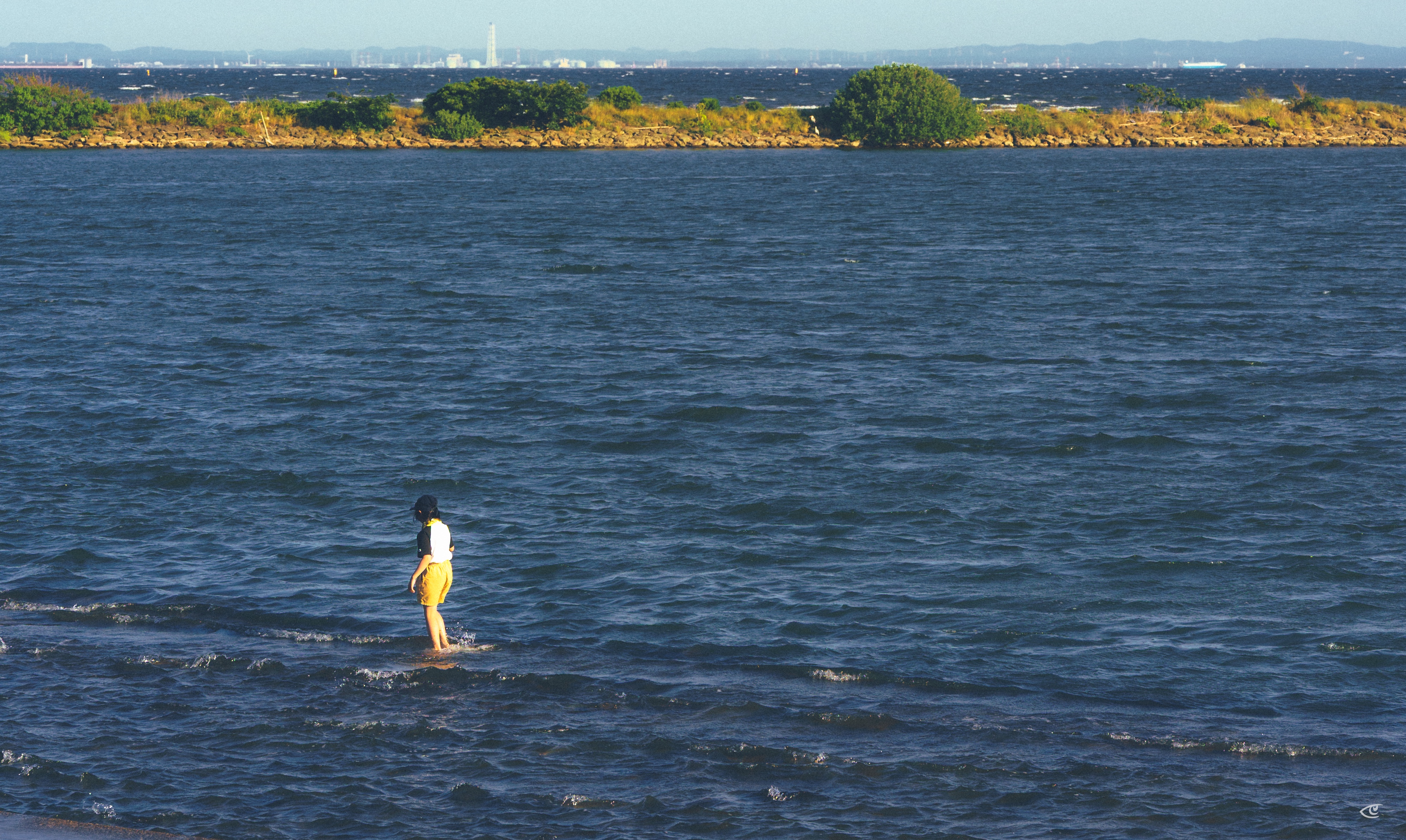 Person wading in shallow, rippling water near the shore, with a low tree-covered strip of land and distant hills on the horizon.