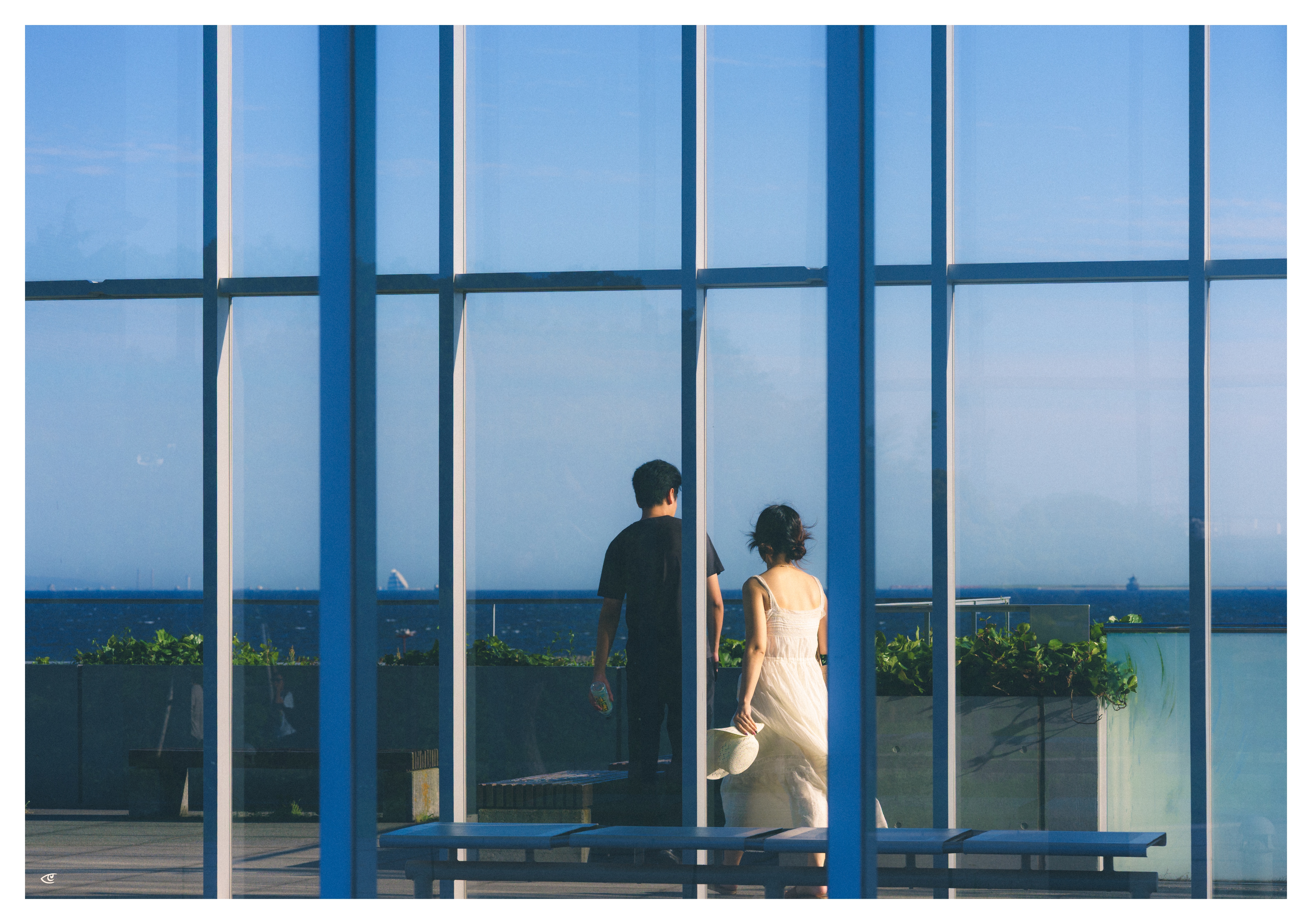 Two people seen through tall blue-framed glass panels, walking on a terrace with planter boxes; one wears dark clothing and the other a light dress, with the ocean and sky visible in the background.