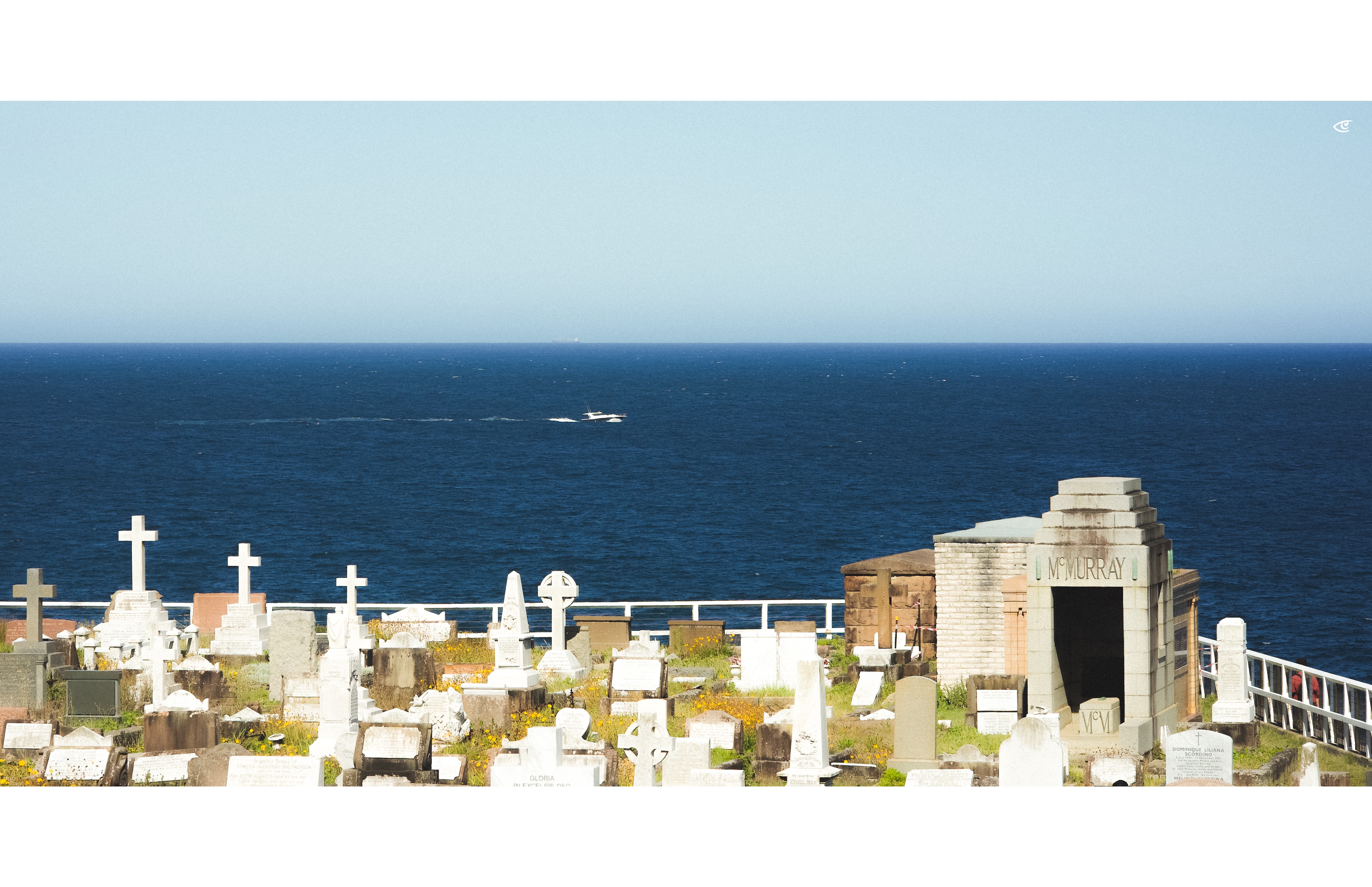 Coastal cemetery with rows of white and gray headstones and crosses in the foreground, a stone mausoleum on the right labeled “M. MURRAY,” and the ocean behind with a small boat leaving a wake near the horizon under a clear sky.