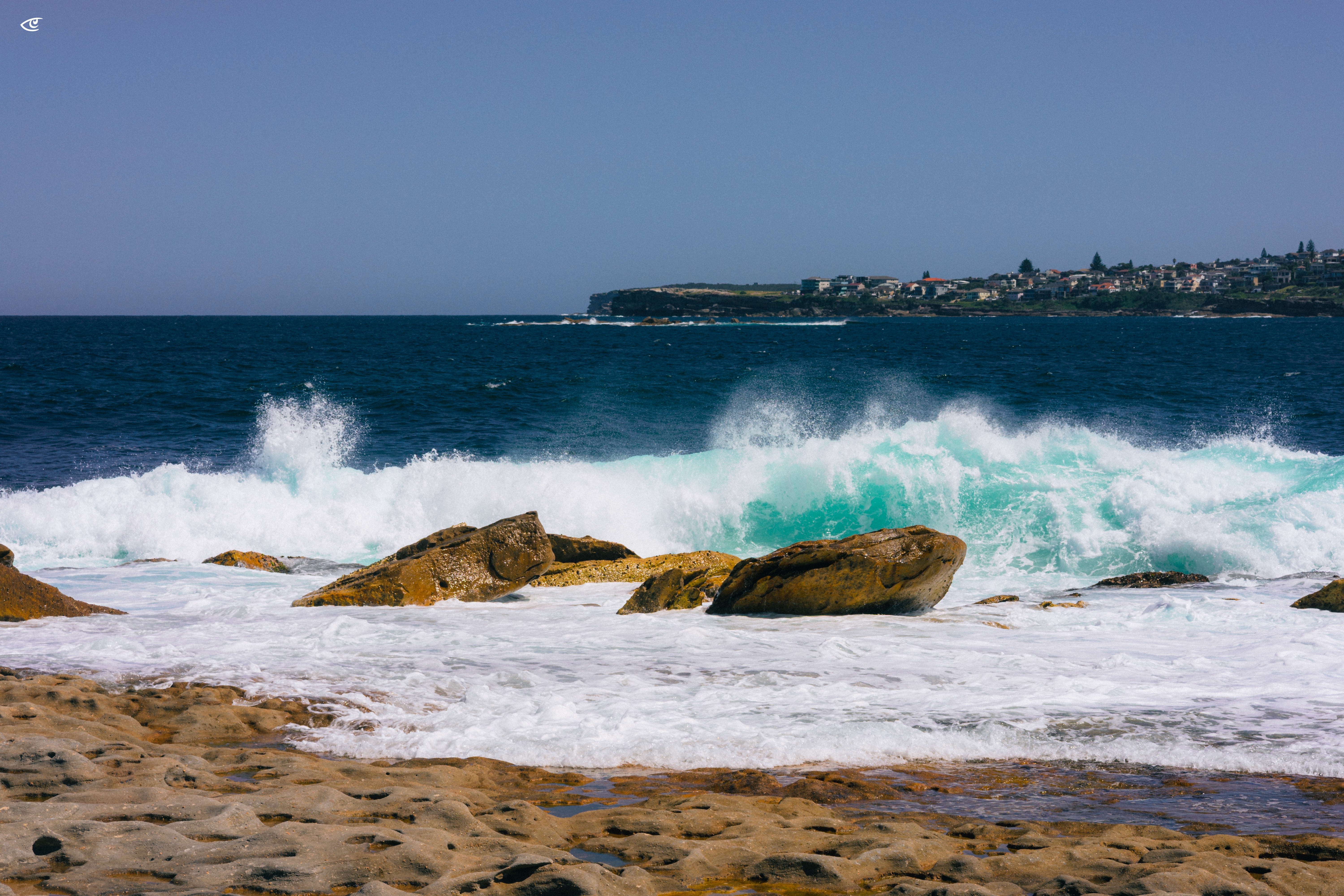 Waves crash over rocky shoreline in the foreground, sending white spray into the air; turquoise surf and deep blue ocean extend to a distant headland with clustered buildings under a clear sky.