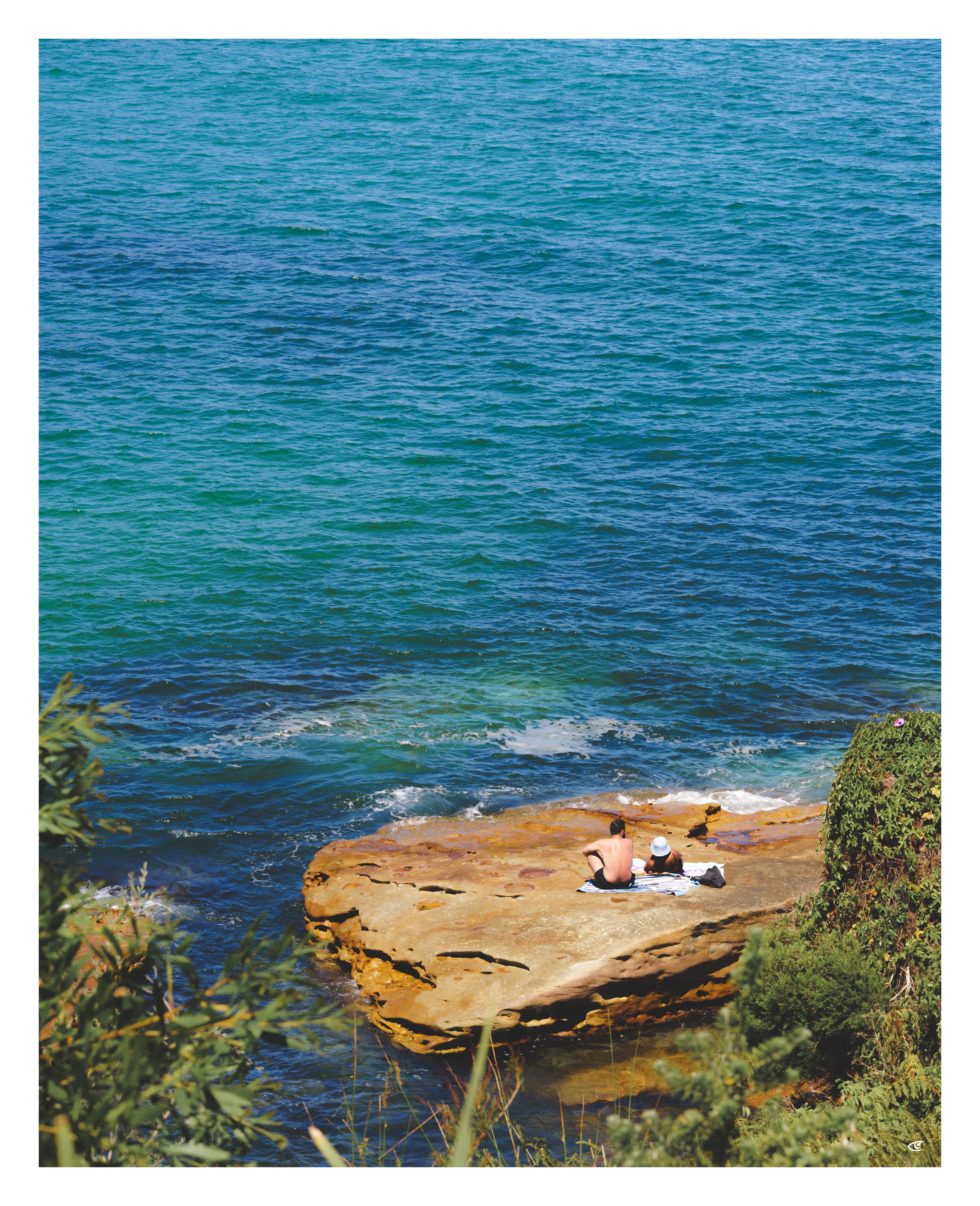 Two people sitting on a towel on a flat rocky ledge by the ocean, with blue-green water filling most of the frame and shrubs in the foreground.