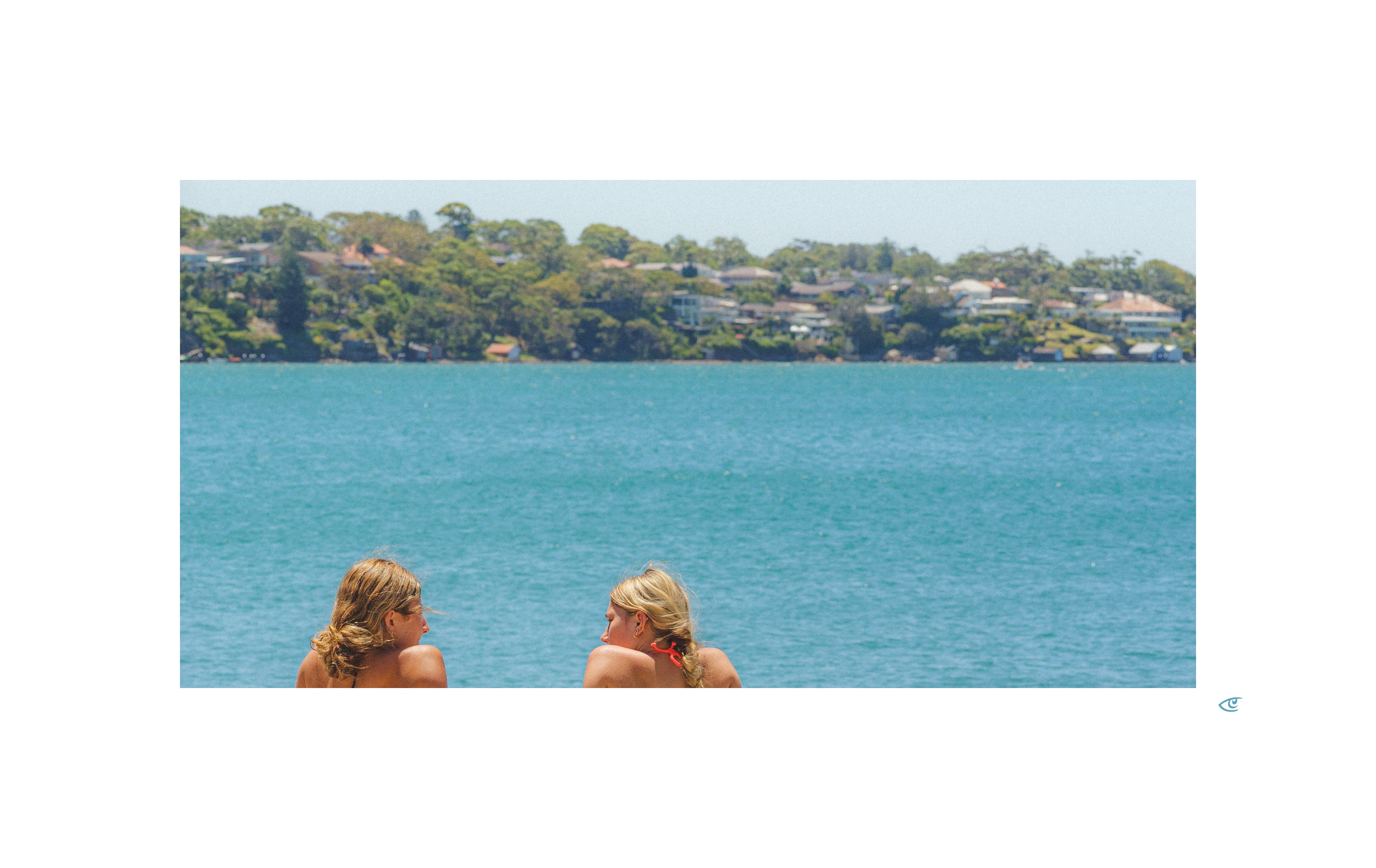 Two women with blonde hair sit in the foreground facing a wide expanse of blue water, with a tree-lined shoreline and scattered houses across the bay under a clear sky.