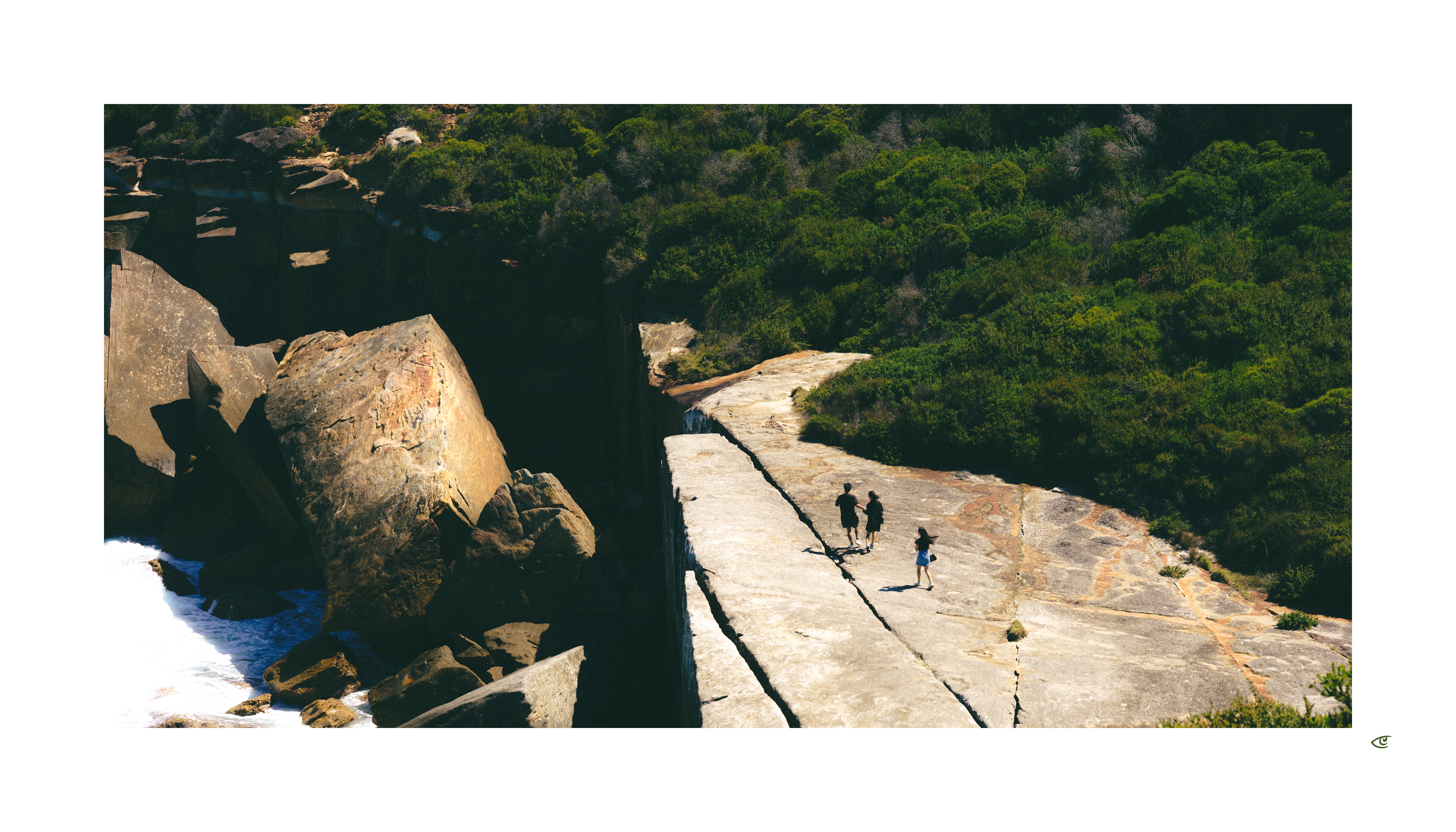 Aerial view of three hikers walking along a wide, pale rock ledge beside a deep, shadowed crevice, with large boulders to the left and dense green vegetation surrounding the rocky area.