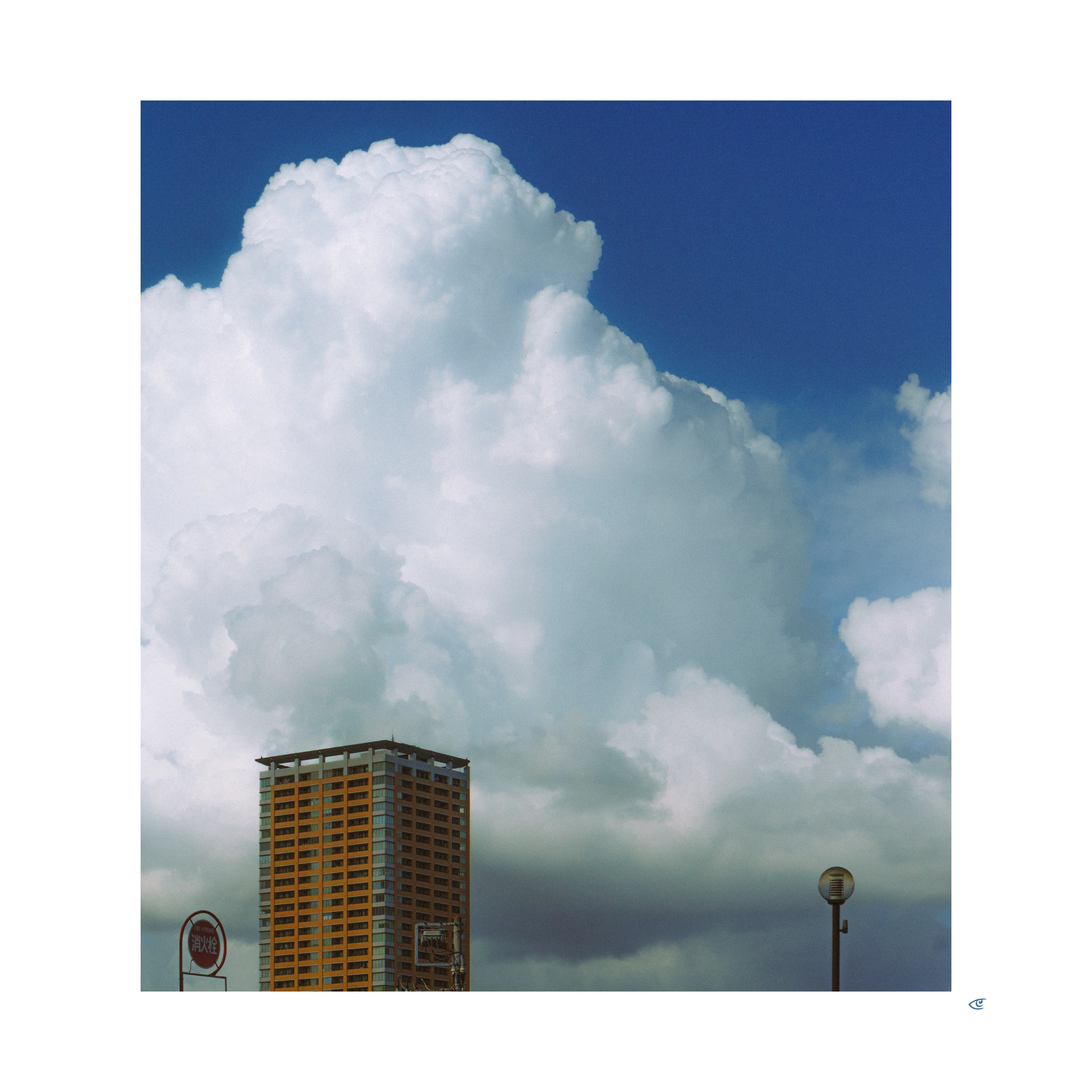 Tall apartment building at the bottom left beneath massive white cumulus clouds against a deep blue sky, with a streetlamp and a small circular sign near the lower edge.