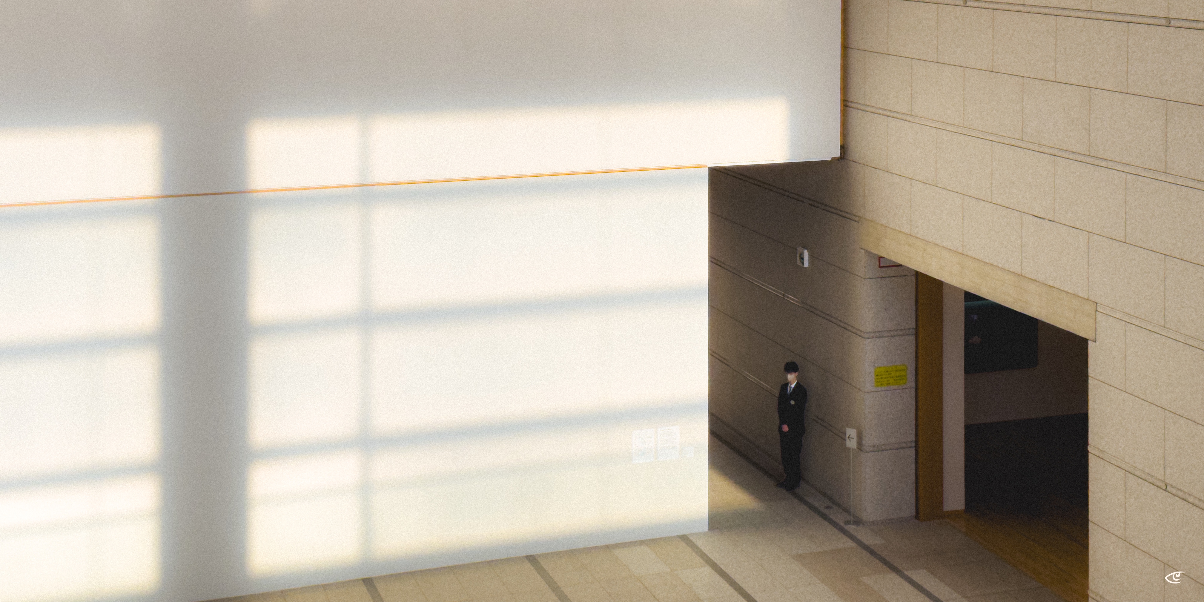 Large white interior wall with sunlight forming a grid of window shadows; at the right, a suited security guard stands beside a doorway in a beige stone corridor.
