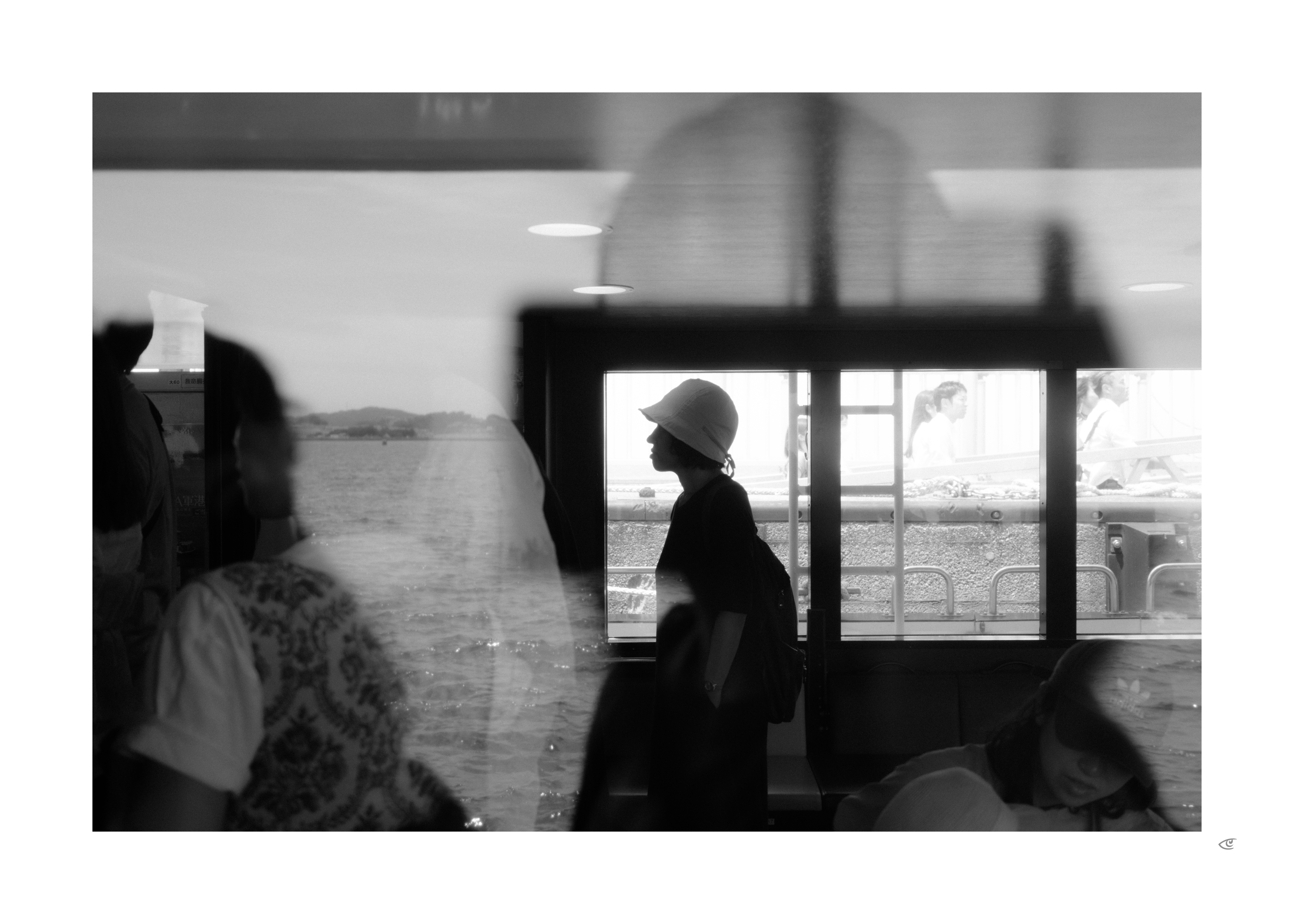 Black-and-white photo with layered reflections: a silhouetted person wearing a brimmed hat and backpack stands in profile by large windows, with water and a distant shoreline visible outside; blurred passengers appear in the foreground, including a seated figure at left and a person with head down at bottom right.
