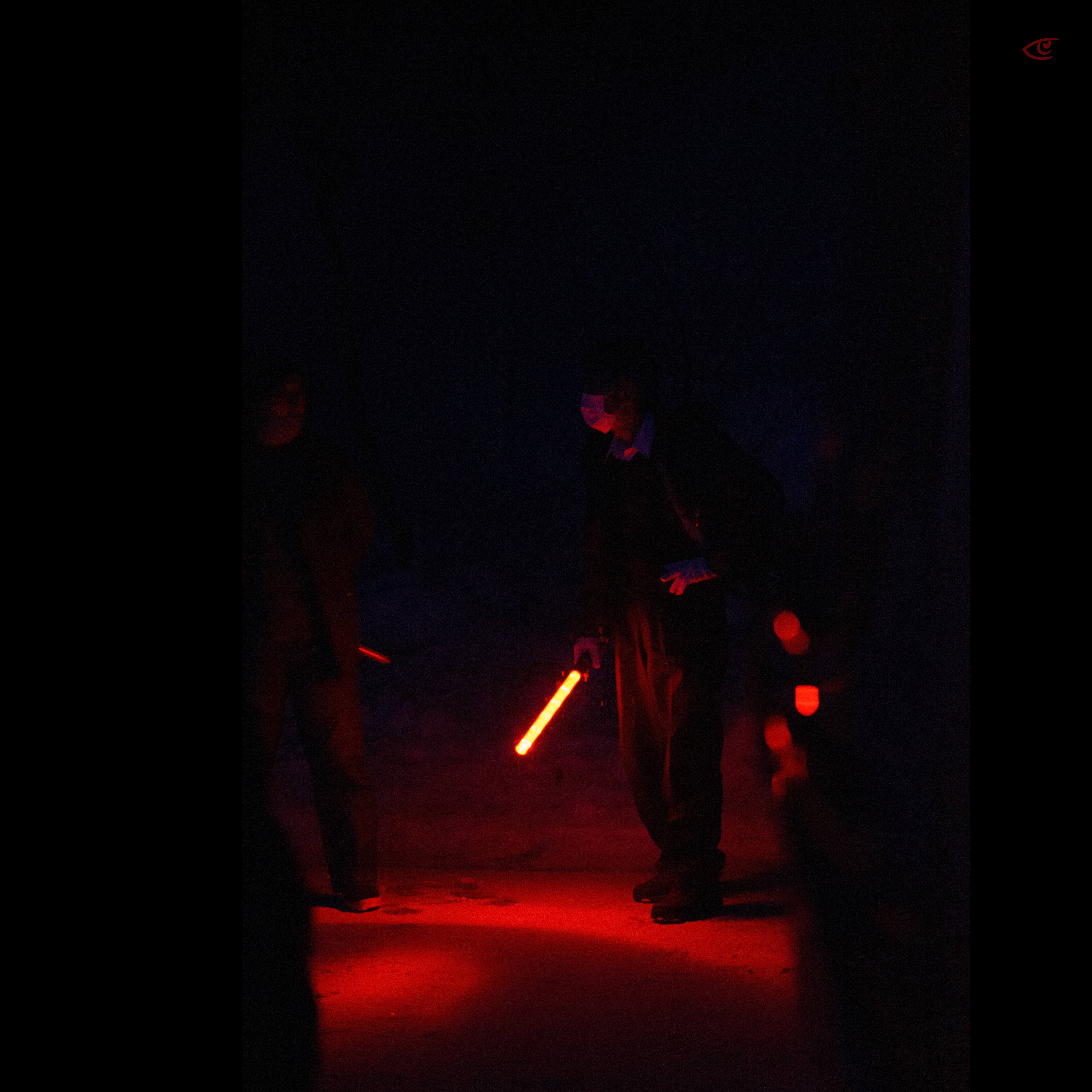 Person standing in a dark, smoky space holding a glowing red light stick pointed downward, casting red light on the ground; another figure partially visible on the left with blurred red lights in the background.
