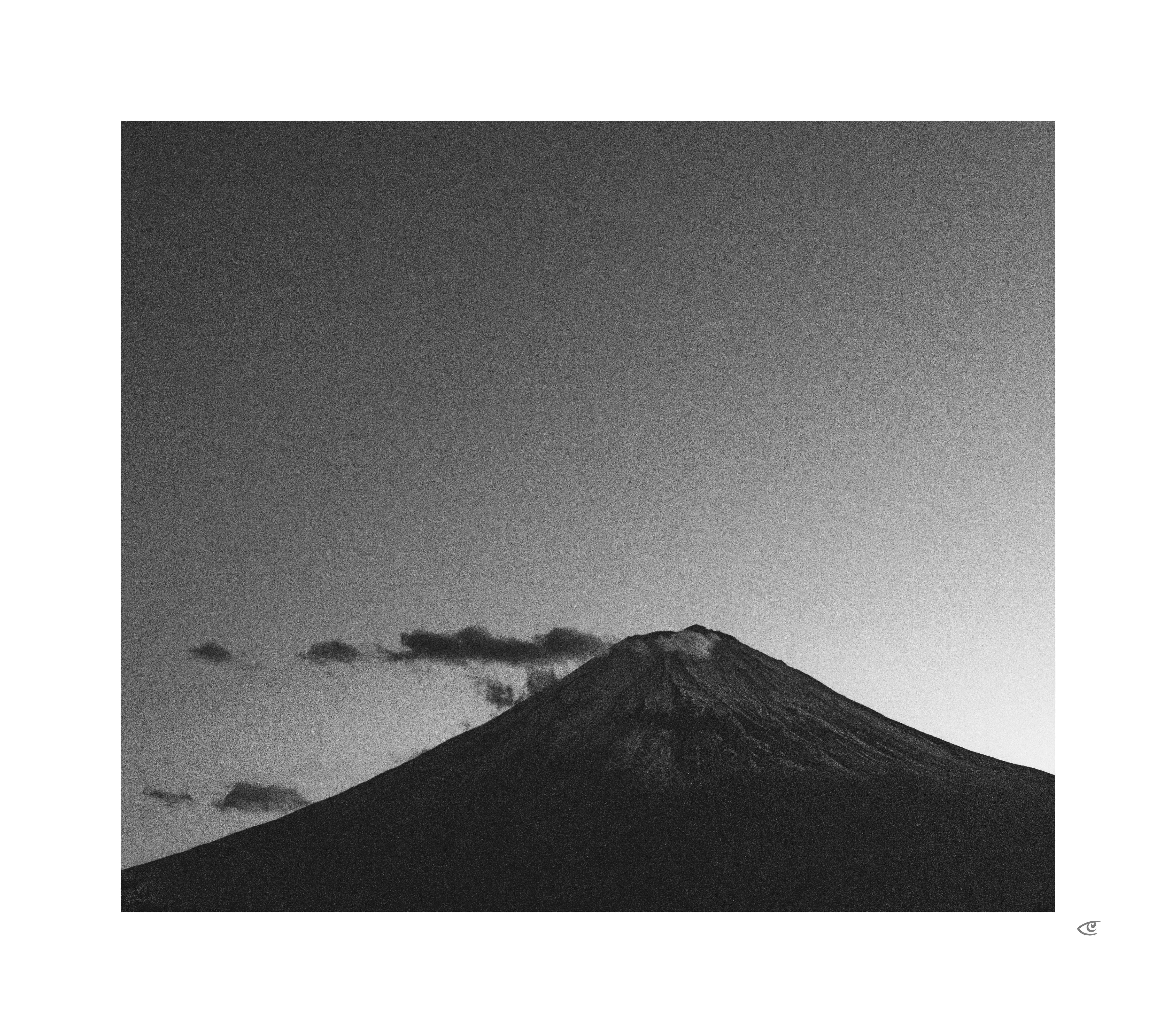 Black-and-white photo of a mountain peak centered low in the frame, with a smooth dark slope and lighter ridges near the summit; a few small clouds drift across the sky to the left, and a bright gradient in the sky appears on the right.