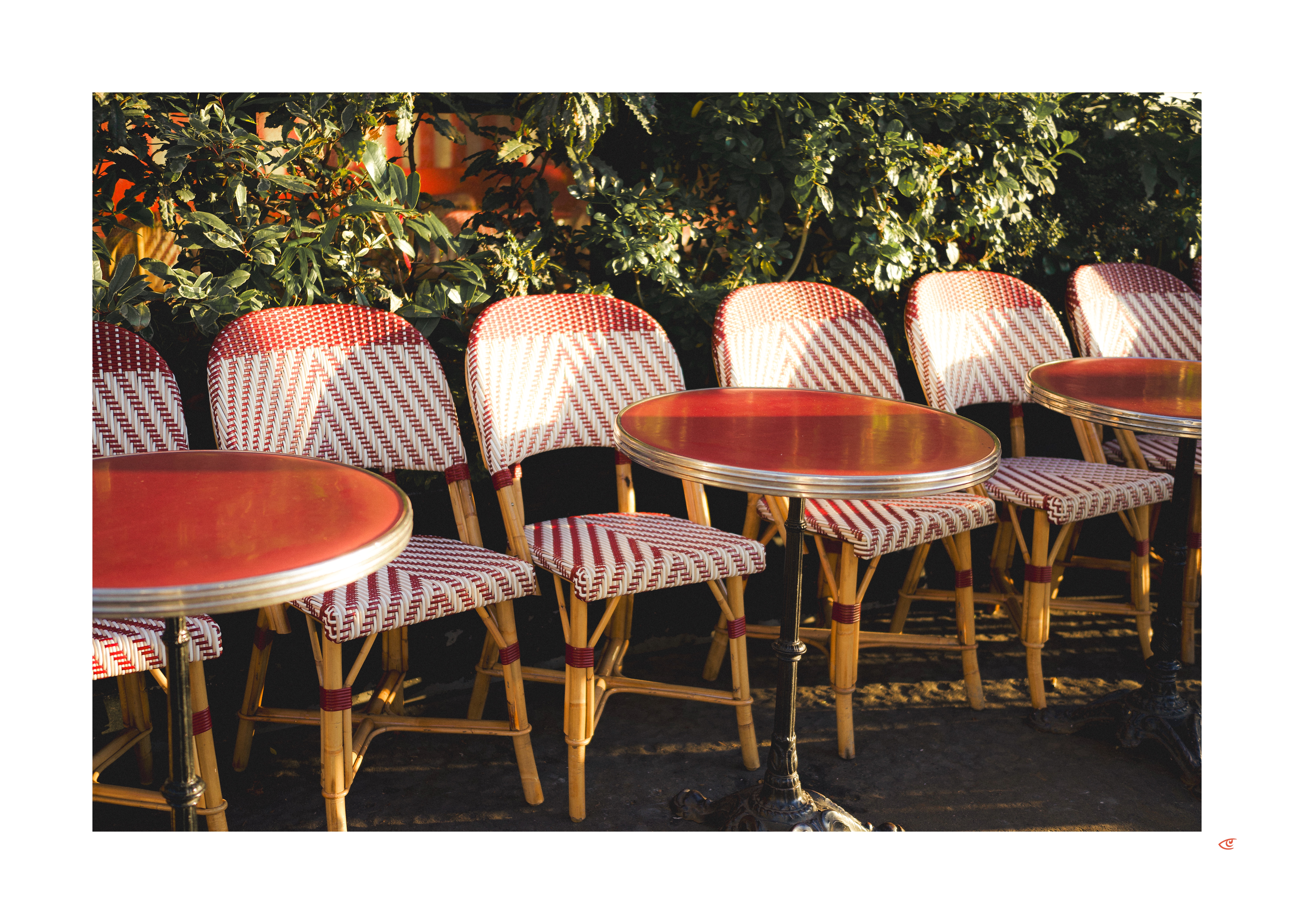 Outdoor café seating with red-and-white woven bistro chairs and two small round tables with red tops, arranged in a row beside dense green shrubs in sunlight.