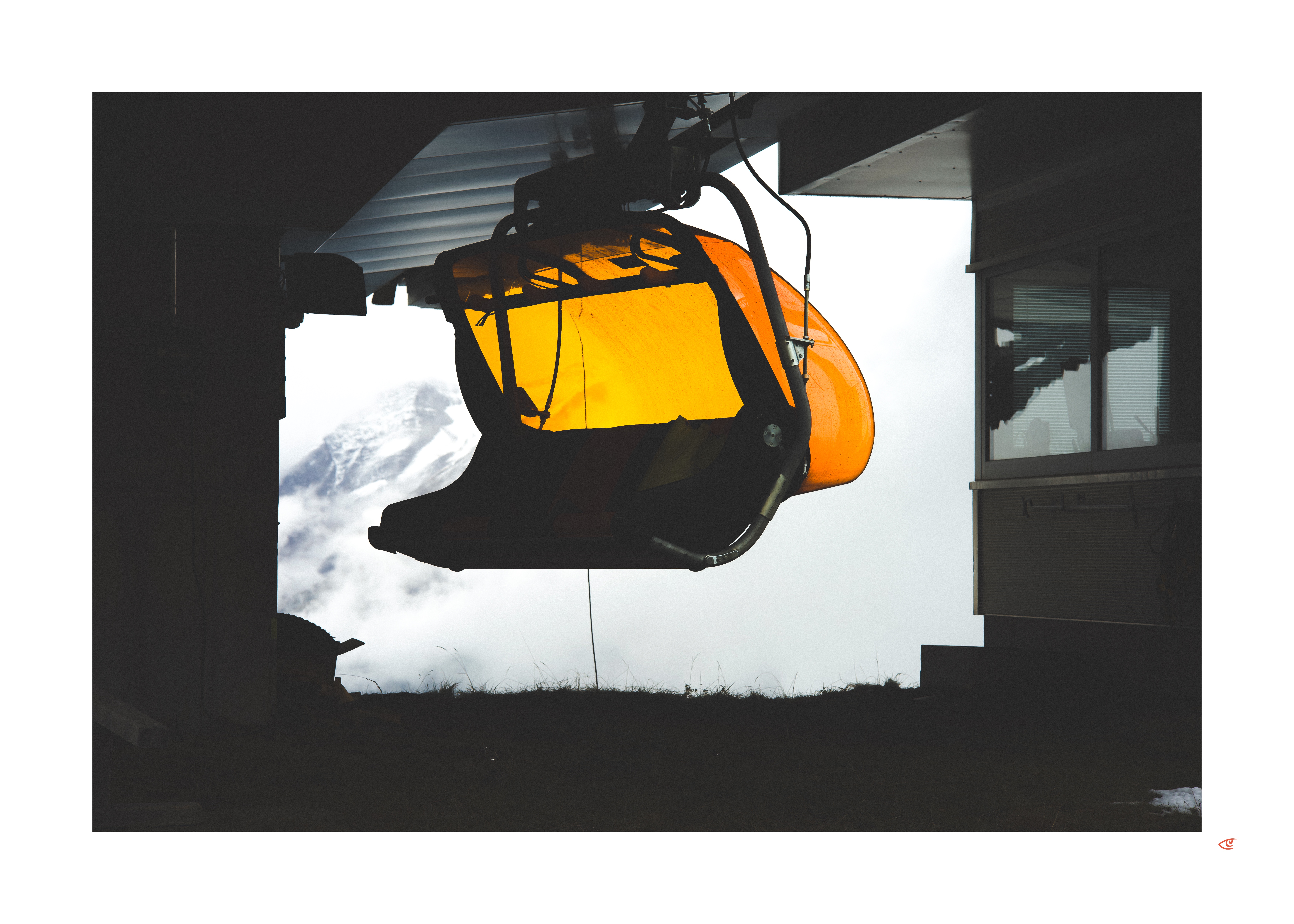 Orange gondola cabin suspended from an overhead cable system inside a dark station, with snowy mountain peaks and clouds visible through the bright opening behind it.
