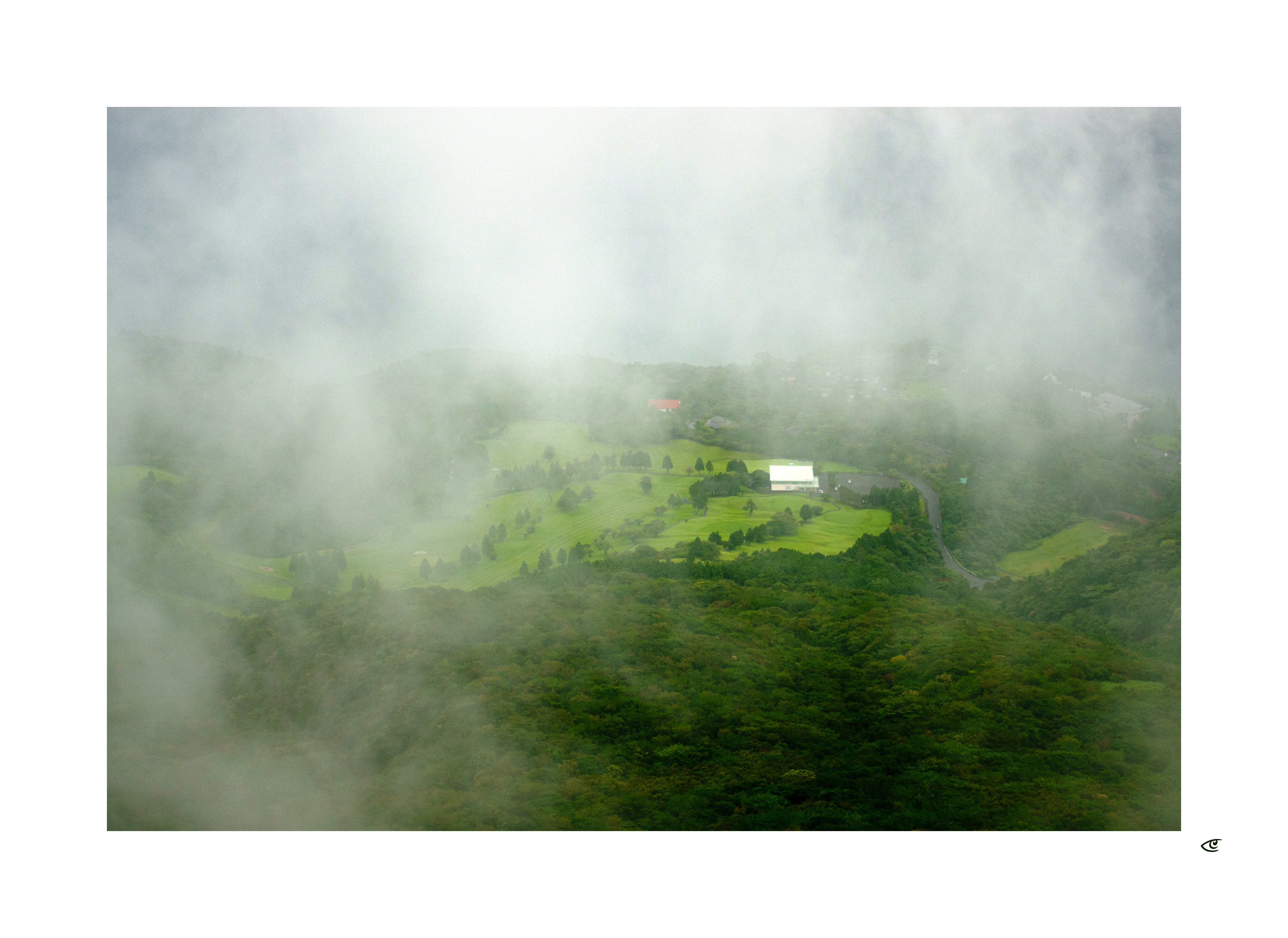 Aerial view of green hills and patchwork fields partly obscured by low clouds, with a winding road and a white-roofed building near the center.