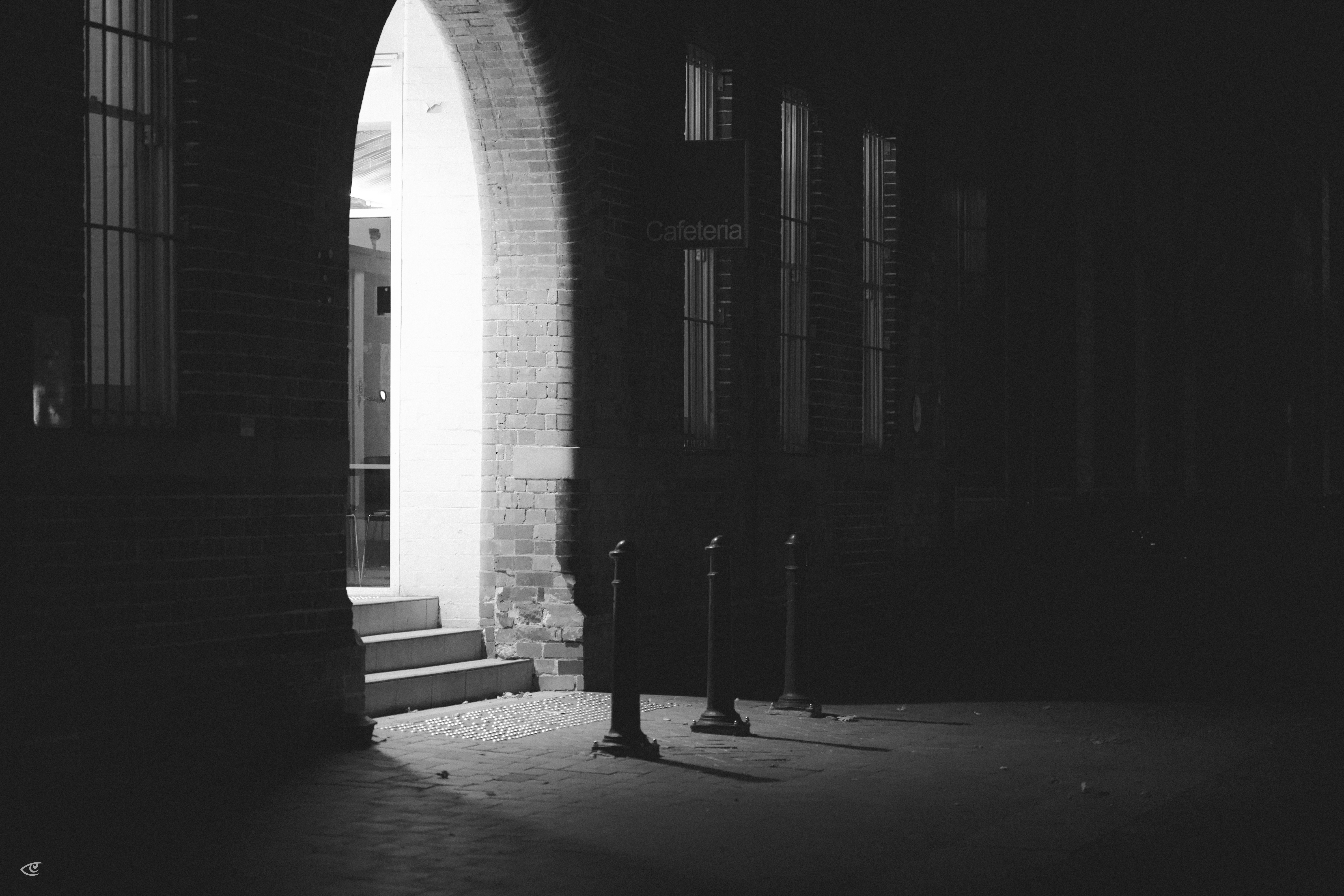 Brick archway leading to a brightly lit interior with a short set of steps; dark exterior corridor in the foreground with several bollards and tall windows along the right wall.