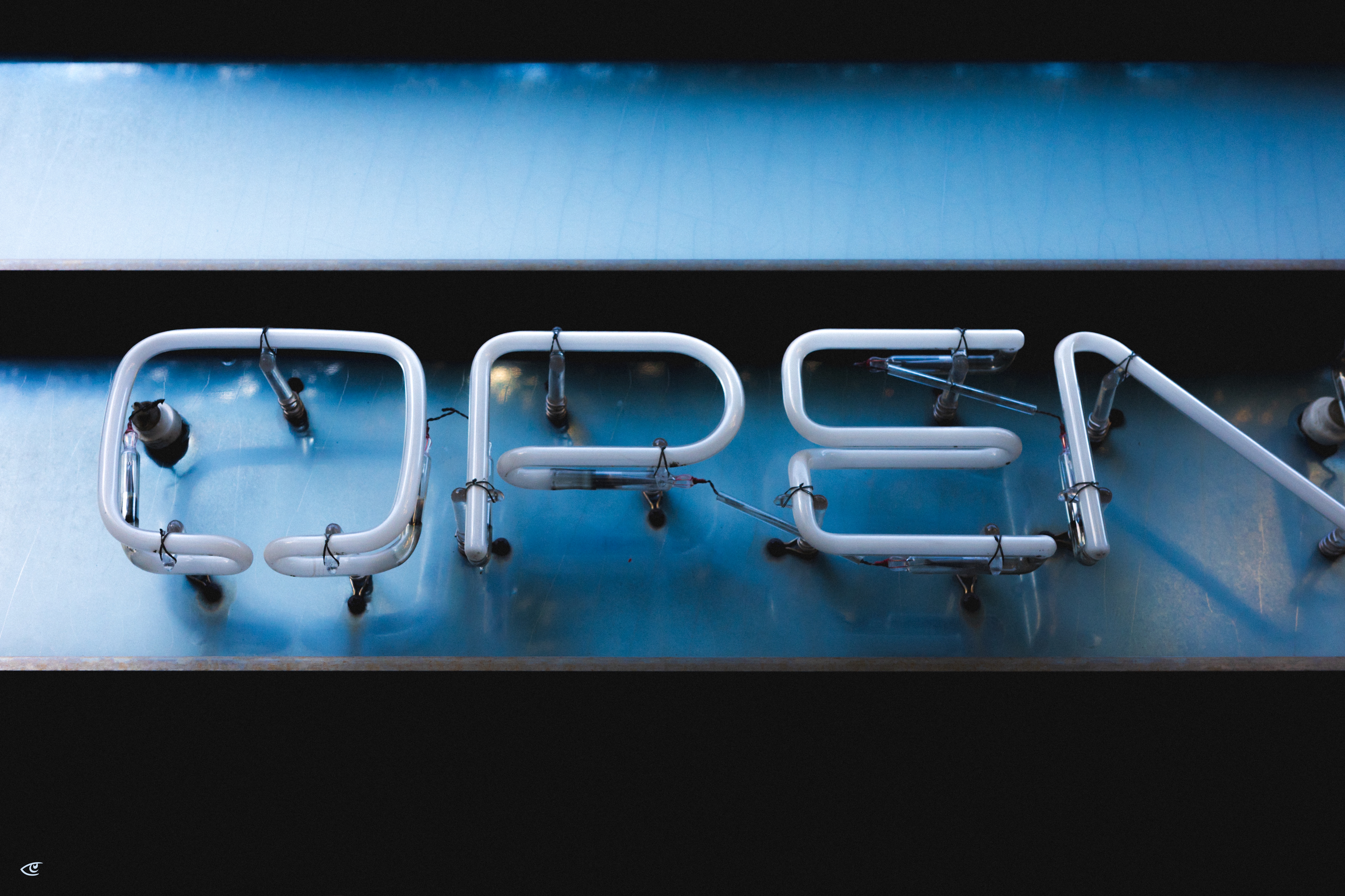 White neon tubing spelling “OPEN” mounted on a blue metal backing, with visible clips and wiring; the right side shows a partially detached tube angled upward.