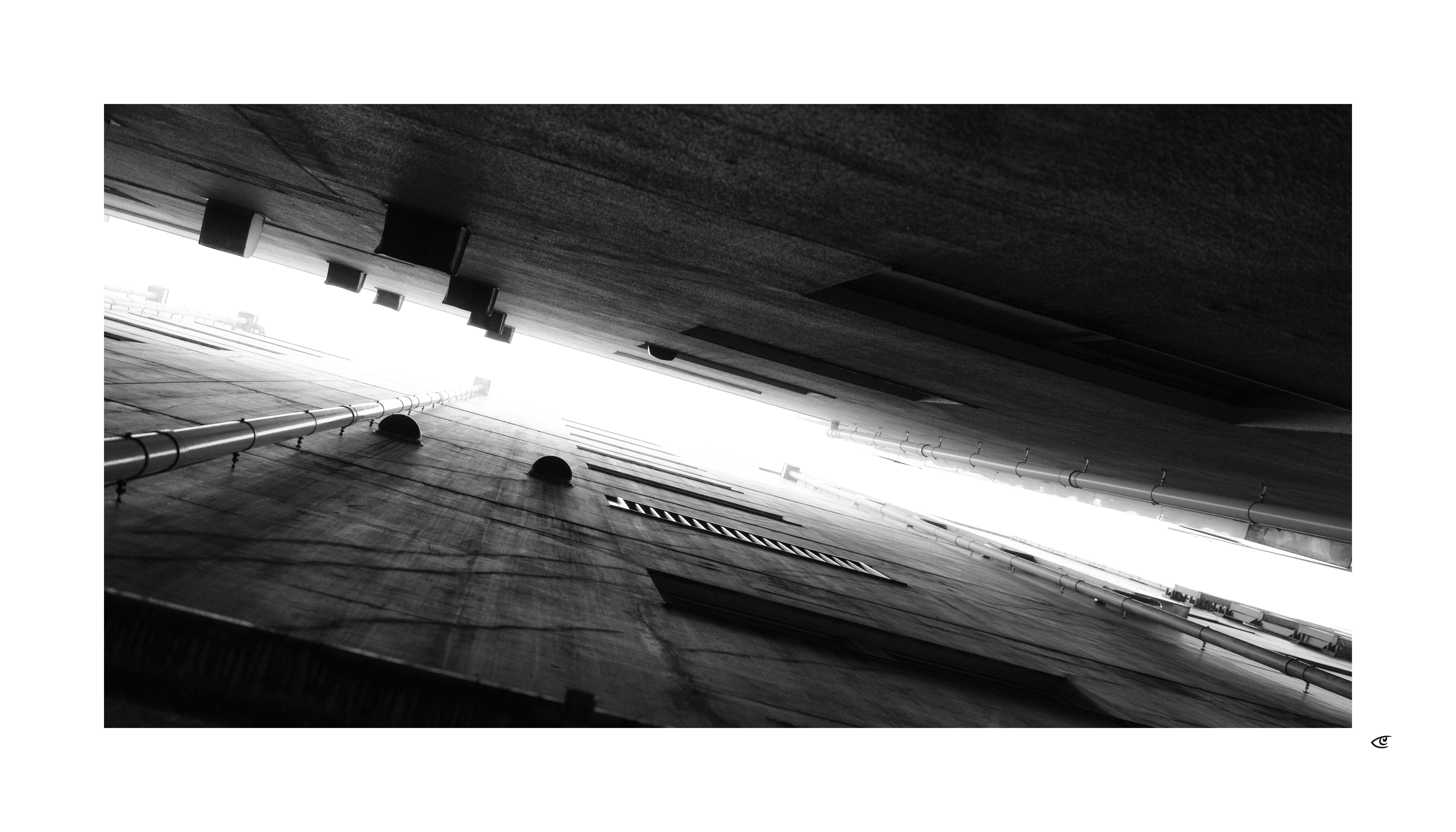 Upward-looking black-and-white view between two tall building walls, with pipes and vents along the surfaces and a bright strip of sky running diagonally across the center.