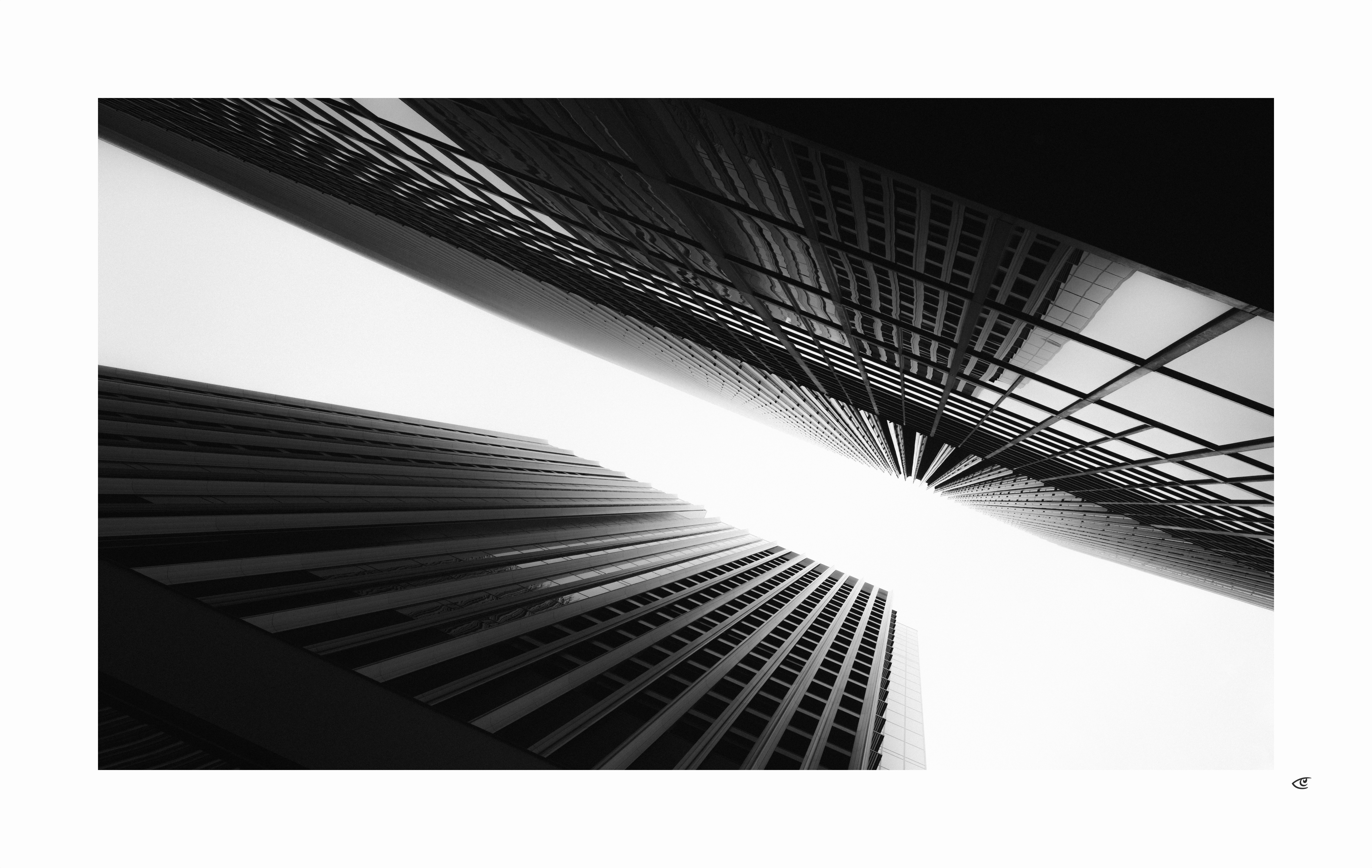 Black-and-white low-angle view of two tall office skyscrapers with repeating window grids, converging toward a bright white sky at the center of the frame.