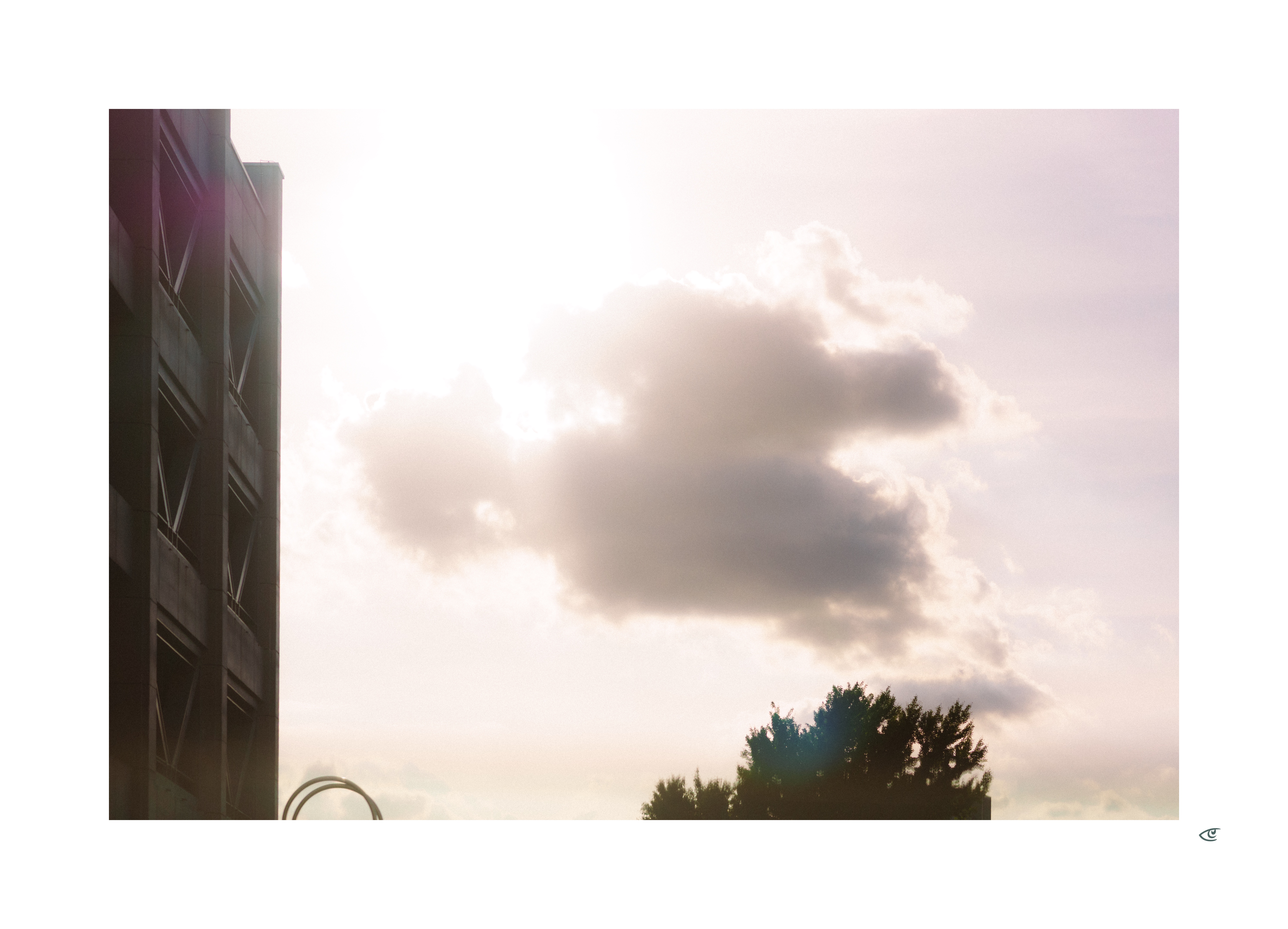 Side view of a tall building along the left edge, with bright sunlit clouds in a pale sky and a line of dark tree tops at the bottom right.