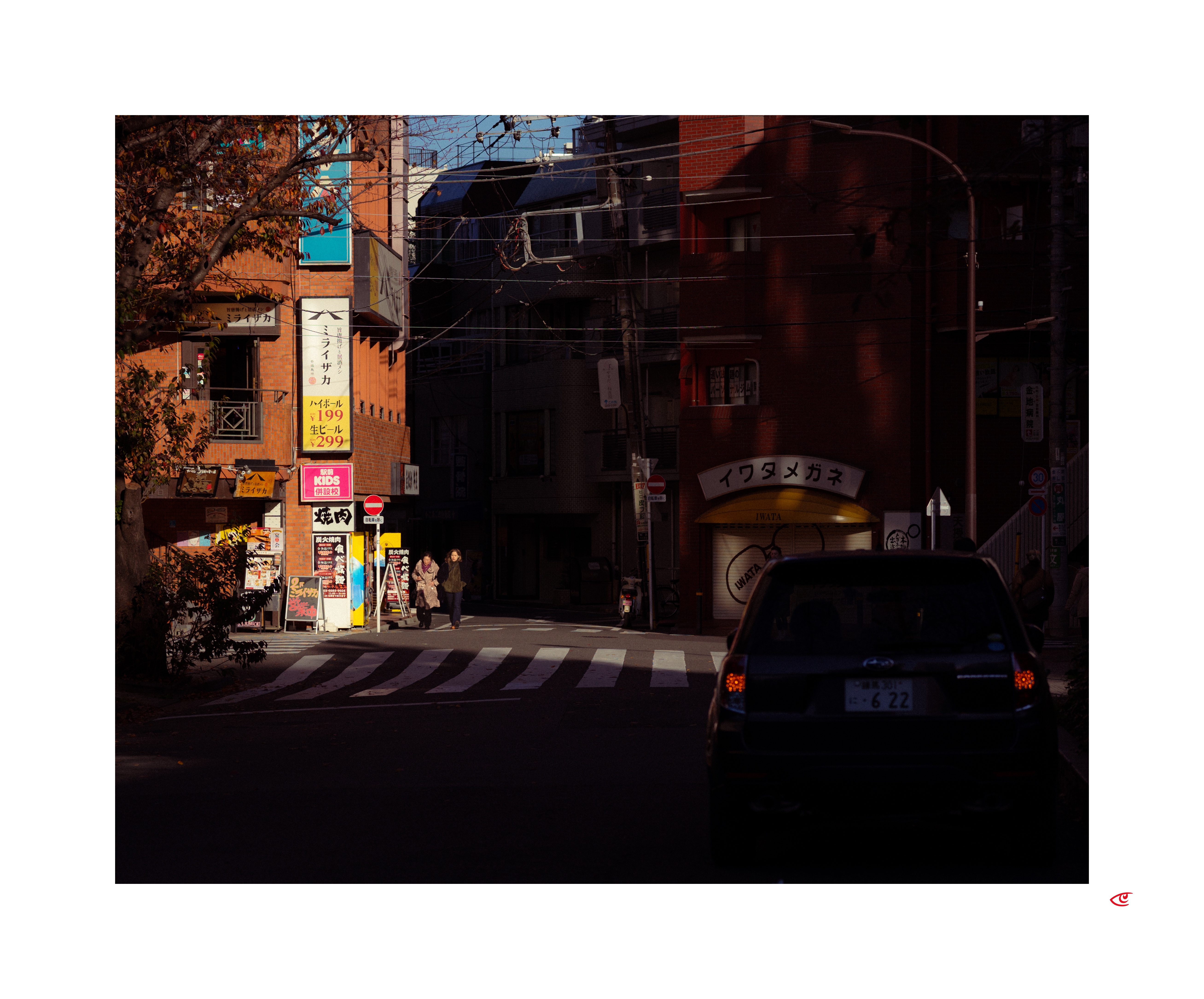 Urban street intersection with a zebra crosswalk, a dark car in the foreground at right, and a few pedestrians near storefronts. Sunlit building facades with Japanese signs on the left contrast with shadowed buildings in the center and right; overhead wires and a tree frame the scene.