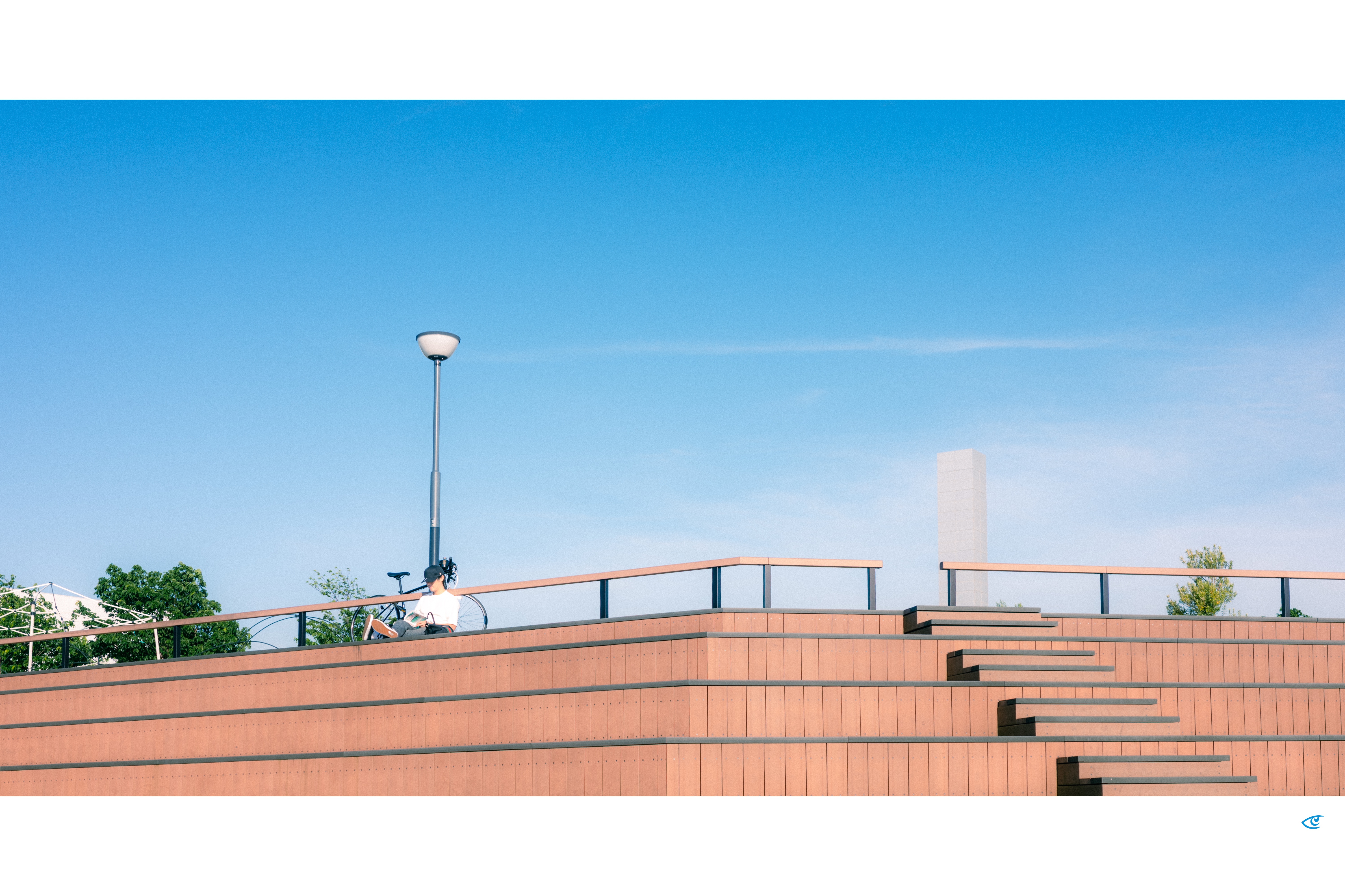Person sitting beside a bicycle on a terraced brick structure with a railing and a tall streetlamp, under a wide blue sky; a stepped staircase rises on the right with a few trees in the background.