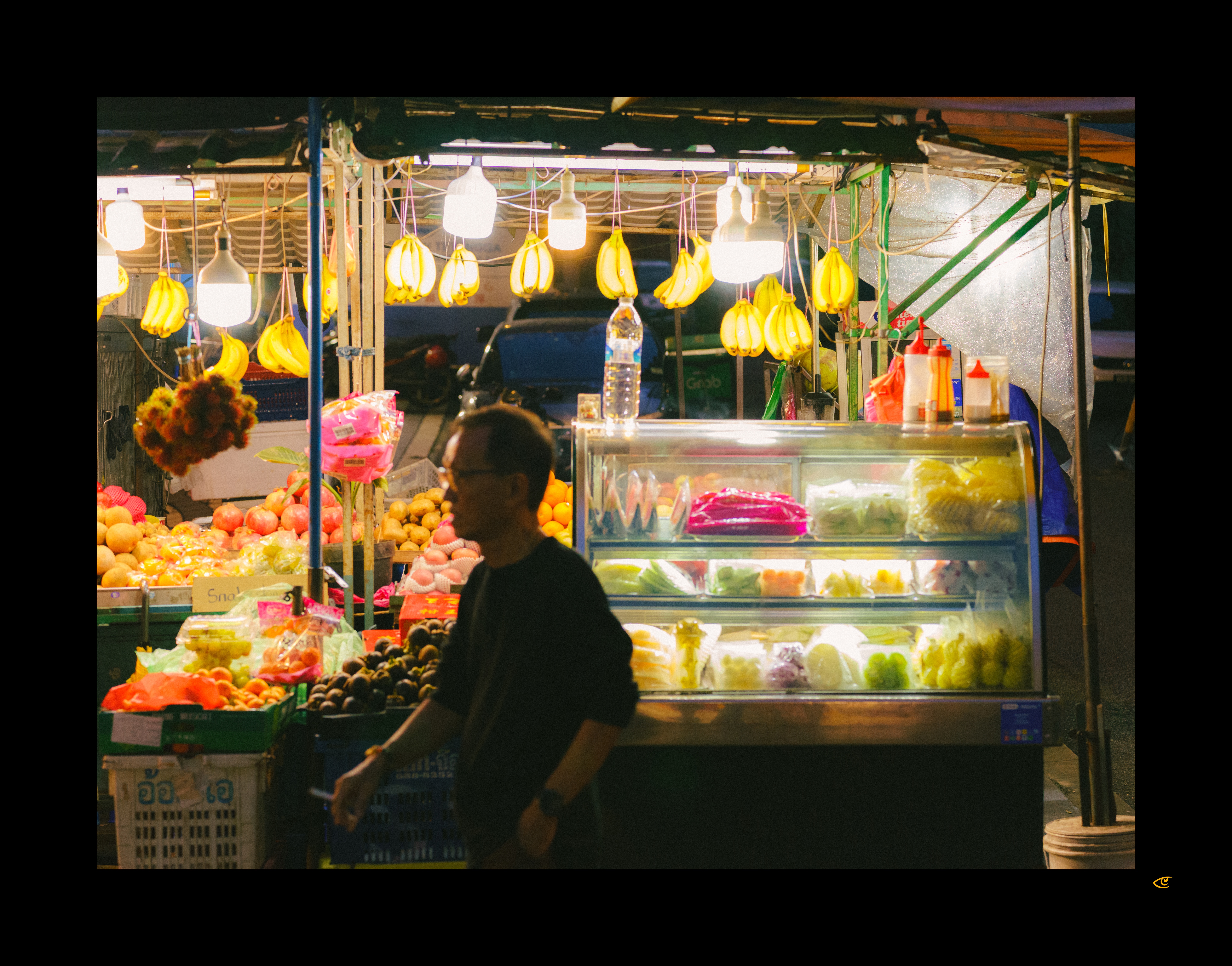 Night street fruit stall with bunches of bananas hanging under bright bulbs, crates of assorted fruit on the left, and a lit glass display case of cut fruit on the right; a man in dark clothing walks past in the foreground.