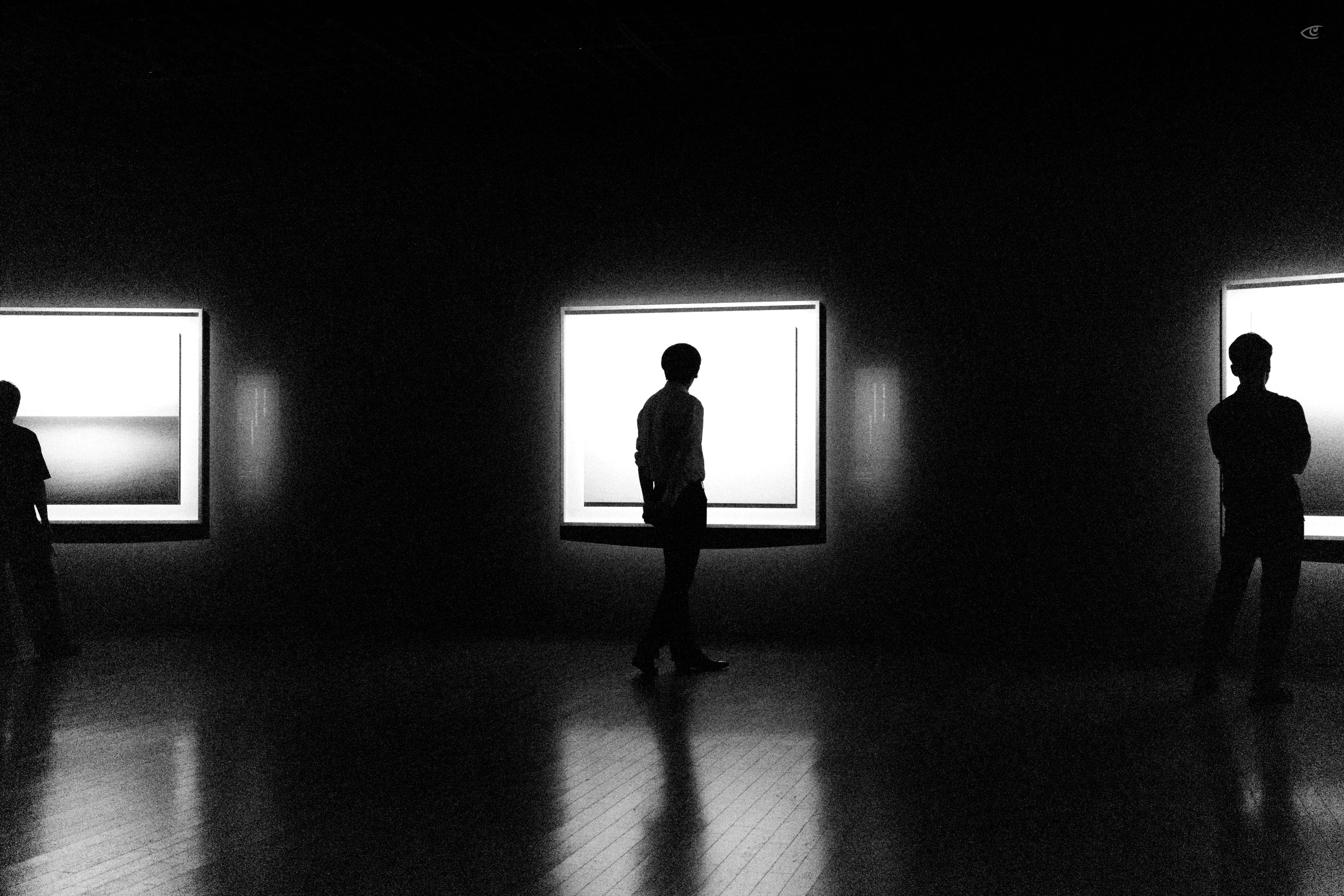 Silhouetted people in a dark gallery stand and walk in front of three large illuminated framed artworks on the wall, with reflections on a polished wooden floor.