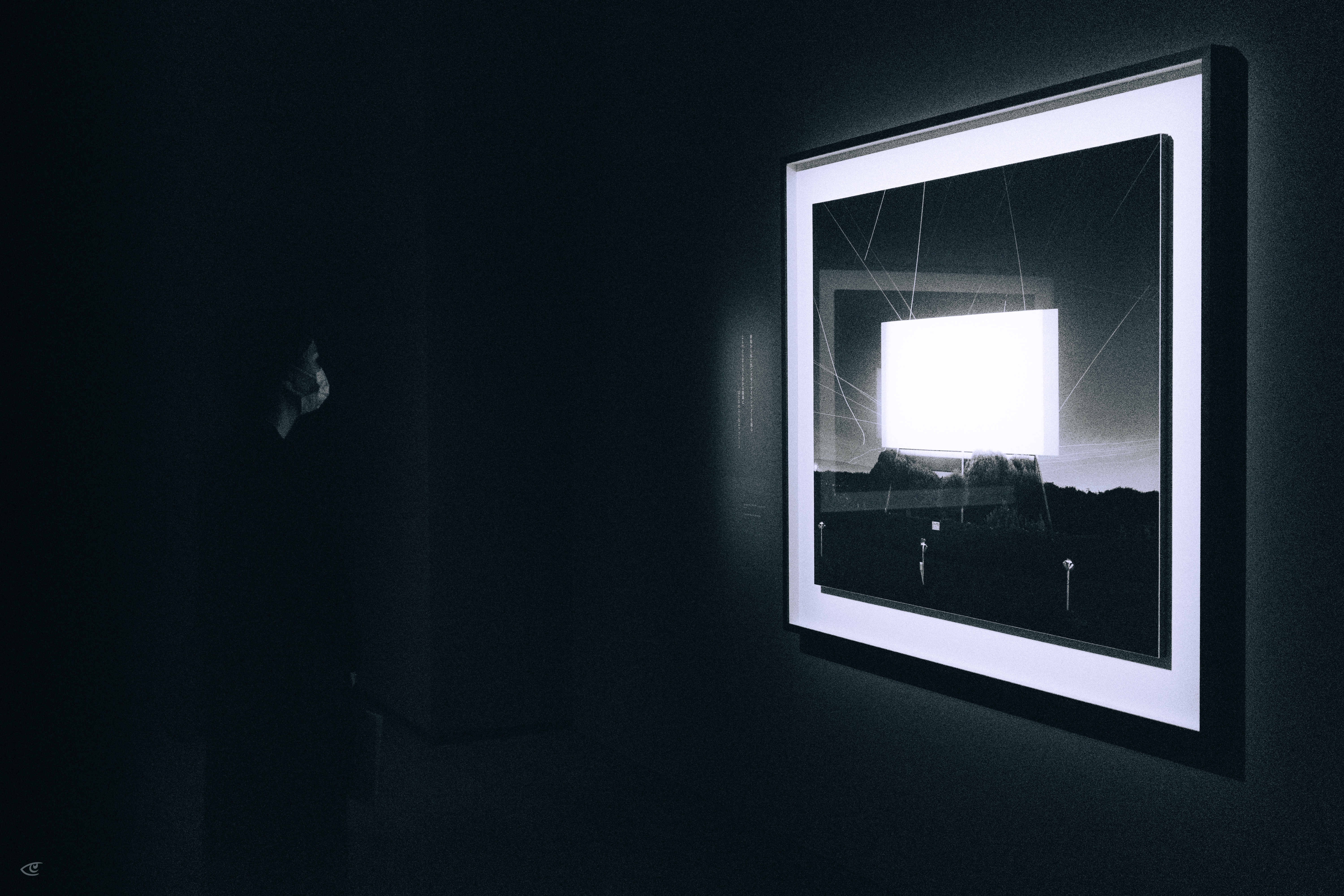 Framed photograph on a dark wall, backlit with a glowing white square in the center; faint silhouette of a person at left facing the artwork.