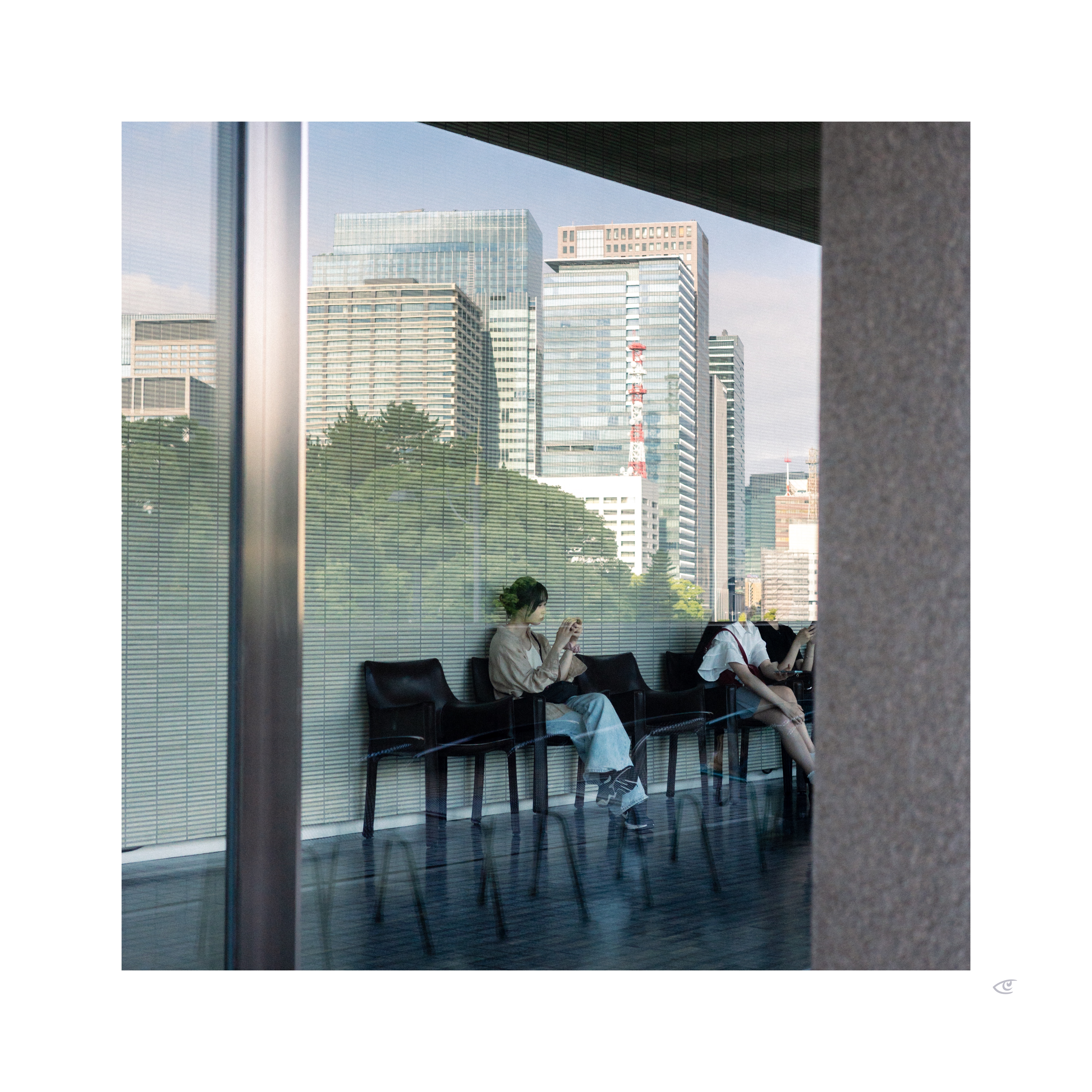 Two people seated on black chairs in a glass-walled lobby, viewed through a reflective window; one looks at a phone while the other leans forward. City skyline with tall office buildings and a red-and-white tower visible outside, with a vertical metal frame and a concrete column bordering the right edge.