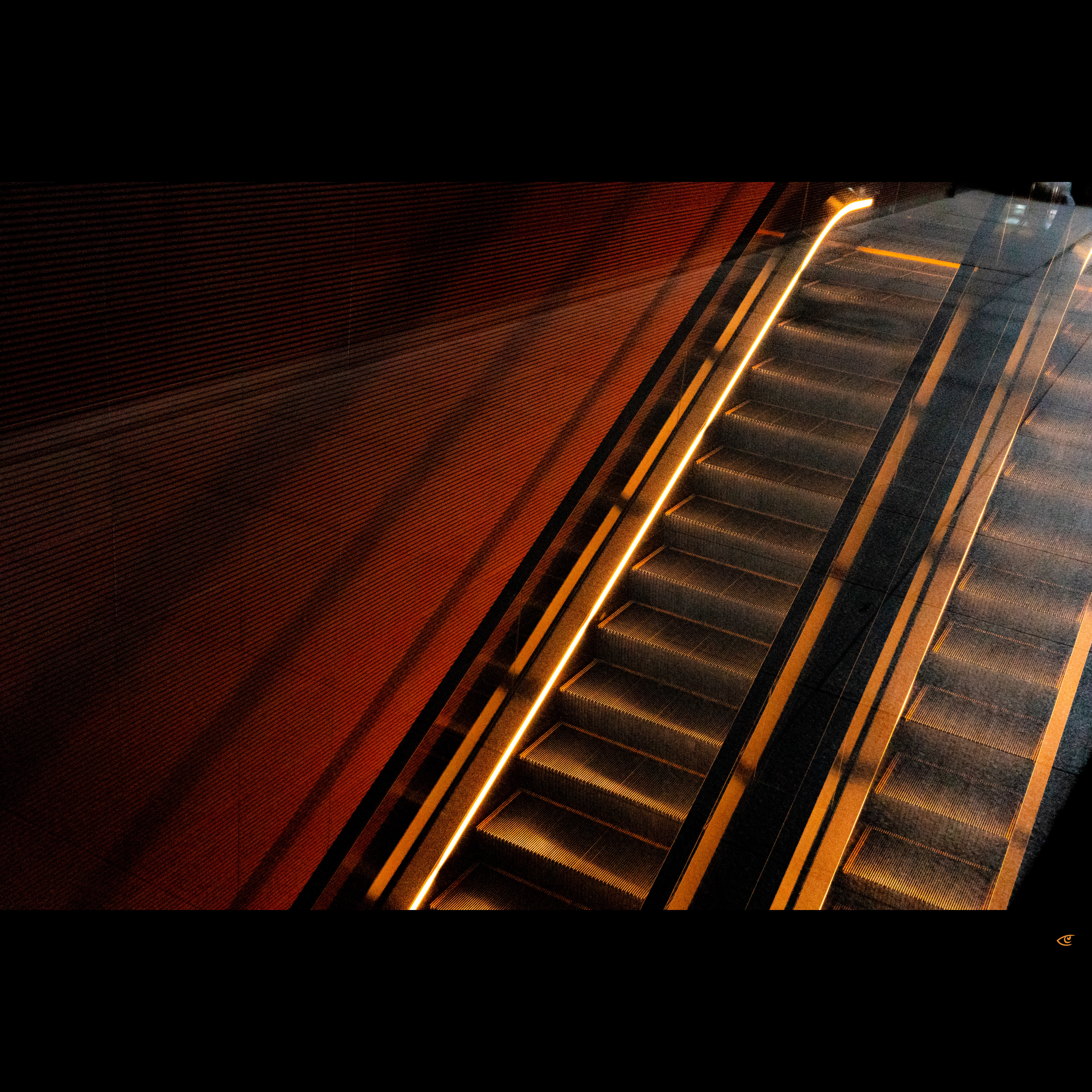 Diagonal overhead view of an escalator with illuminated handrails running upward beside a textured orange wall, with a second escalator partially visible on the right.