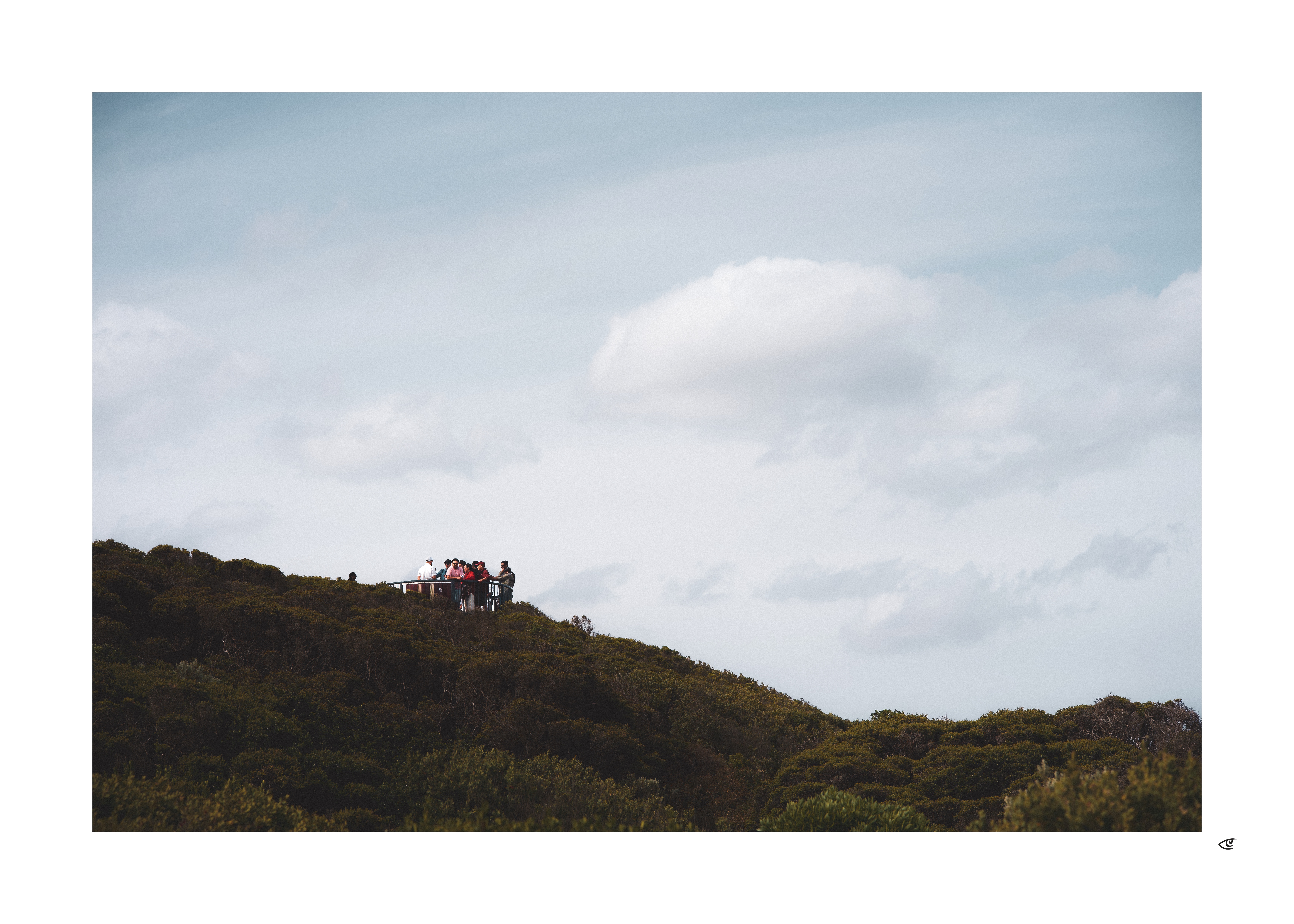 Small group of people standing on a lookout platform on a forested hill, with a wide cloudy sky above.