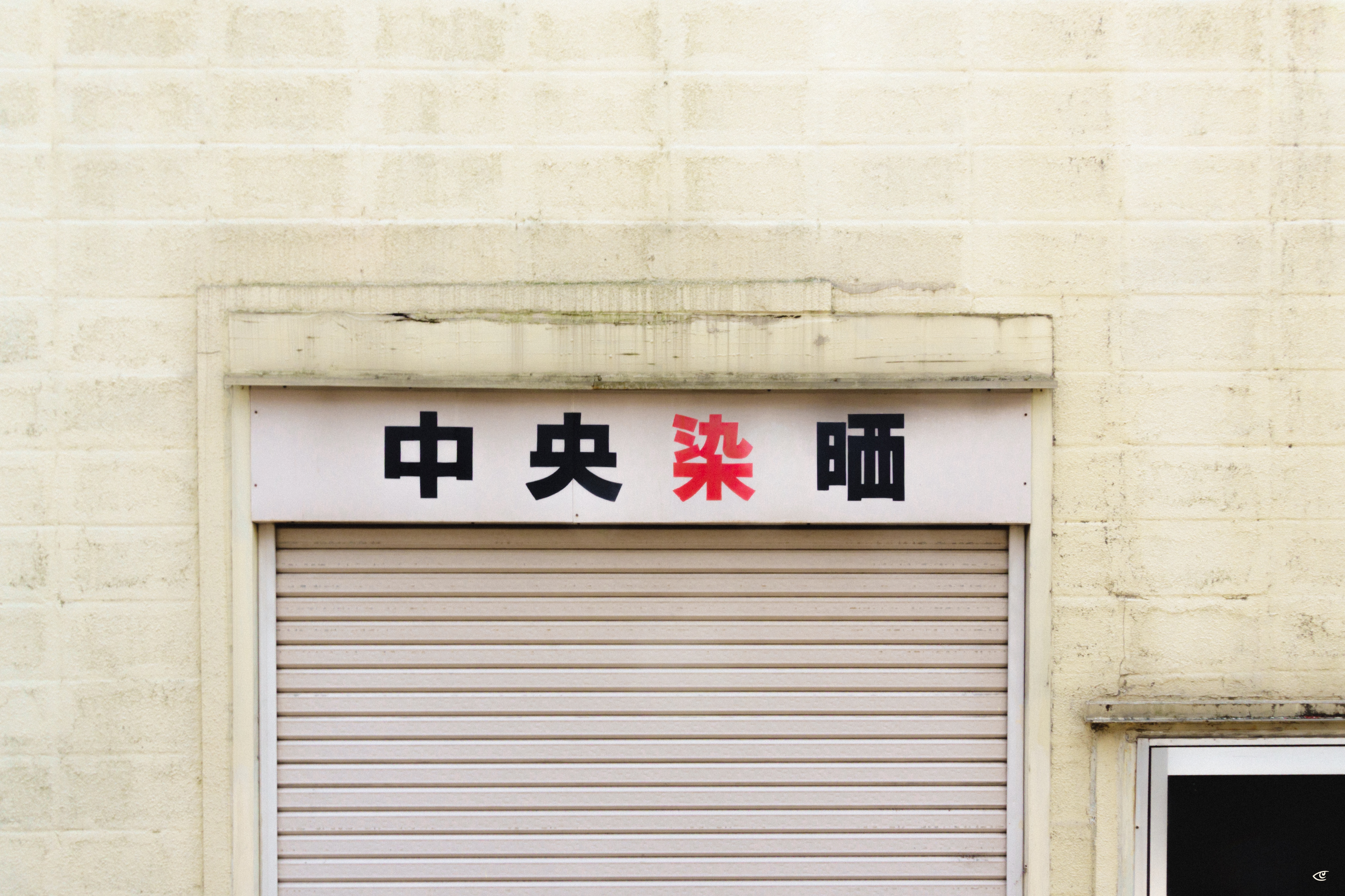 White exterior wall with a closed gray roll-up shutter; above it, a horizontal sign with four Japanese characters in black and red, and a window frame partially visible on the right.