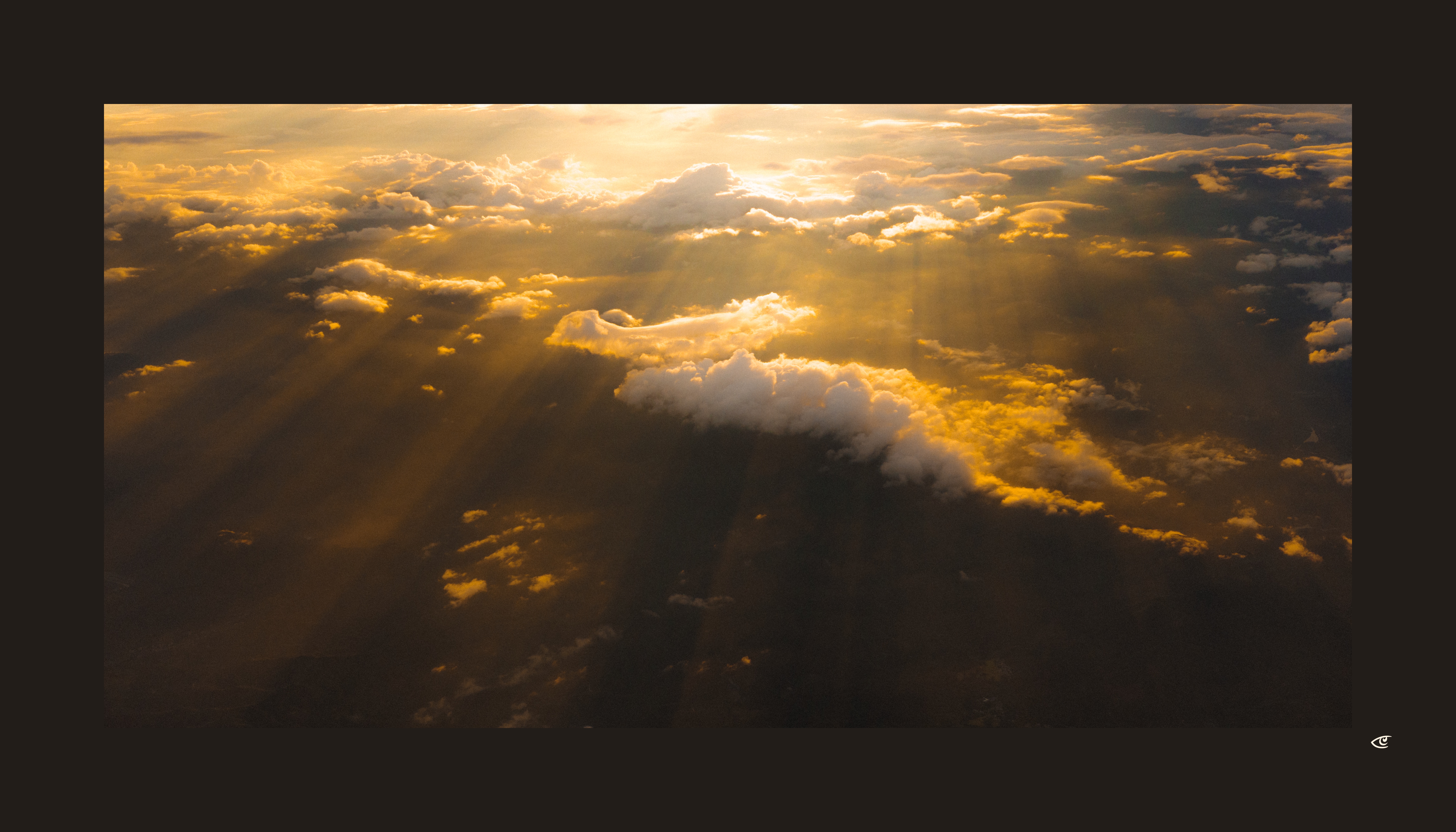 Aerial view of thick clouds lit by low sunlight, with golden rays streaming through gaps and casting long shadows across darker cloud layers below.