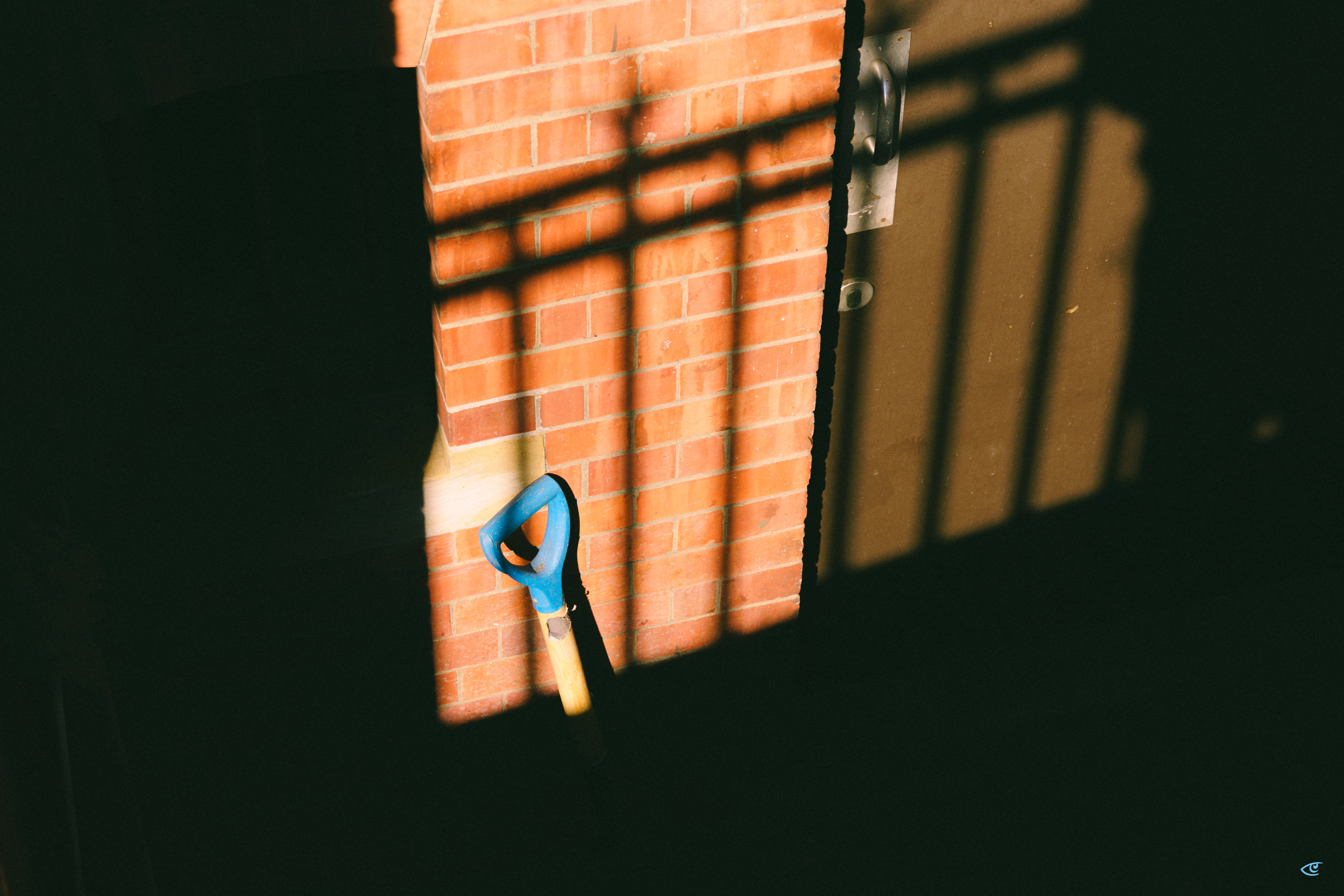 Blue-handled shovel leaning against a red brick wall beside a dark door, with strong sunlight and window-grid shadows cast across the wall and ground.