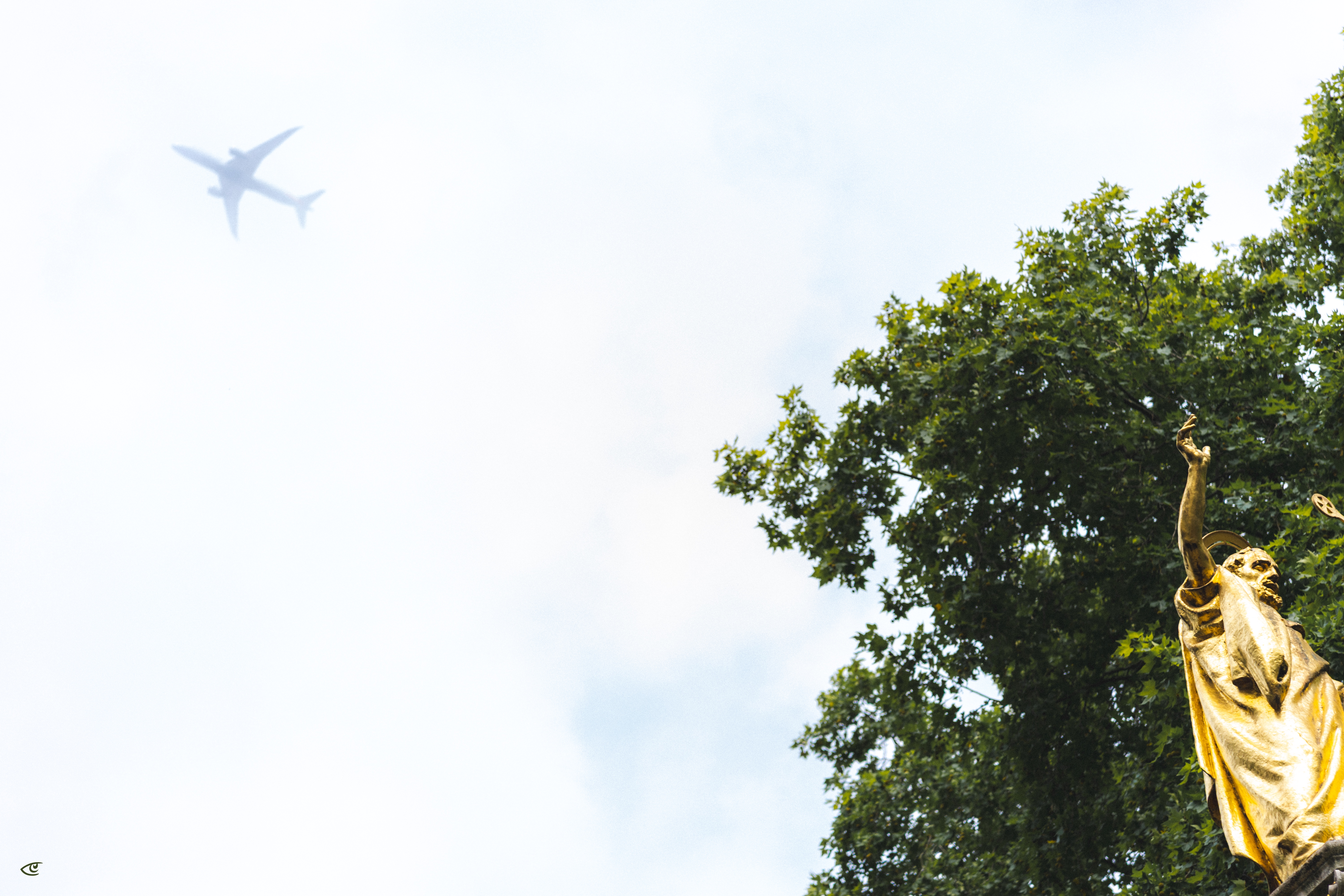Distant airplane silhouette in a bright, cloudy sky above leafy green treetops, with a gold statue on the right raising one arm.