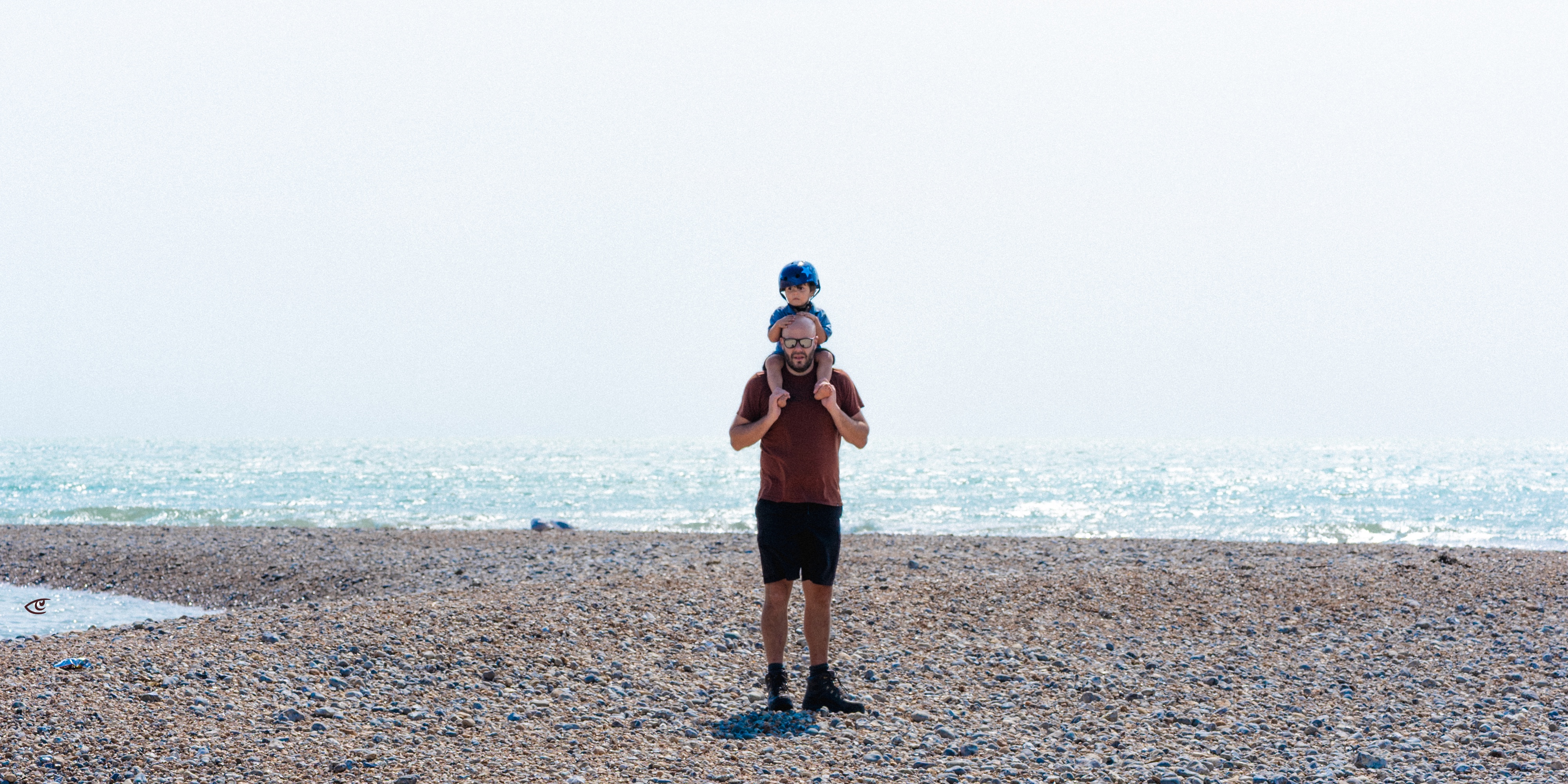 Adult standing on a pebbled beach with a child sitting on their shoulders, facing the camera, with the sea and bright sky in the background.