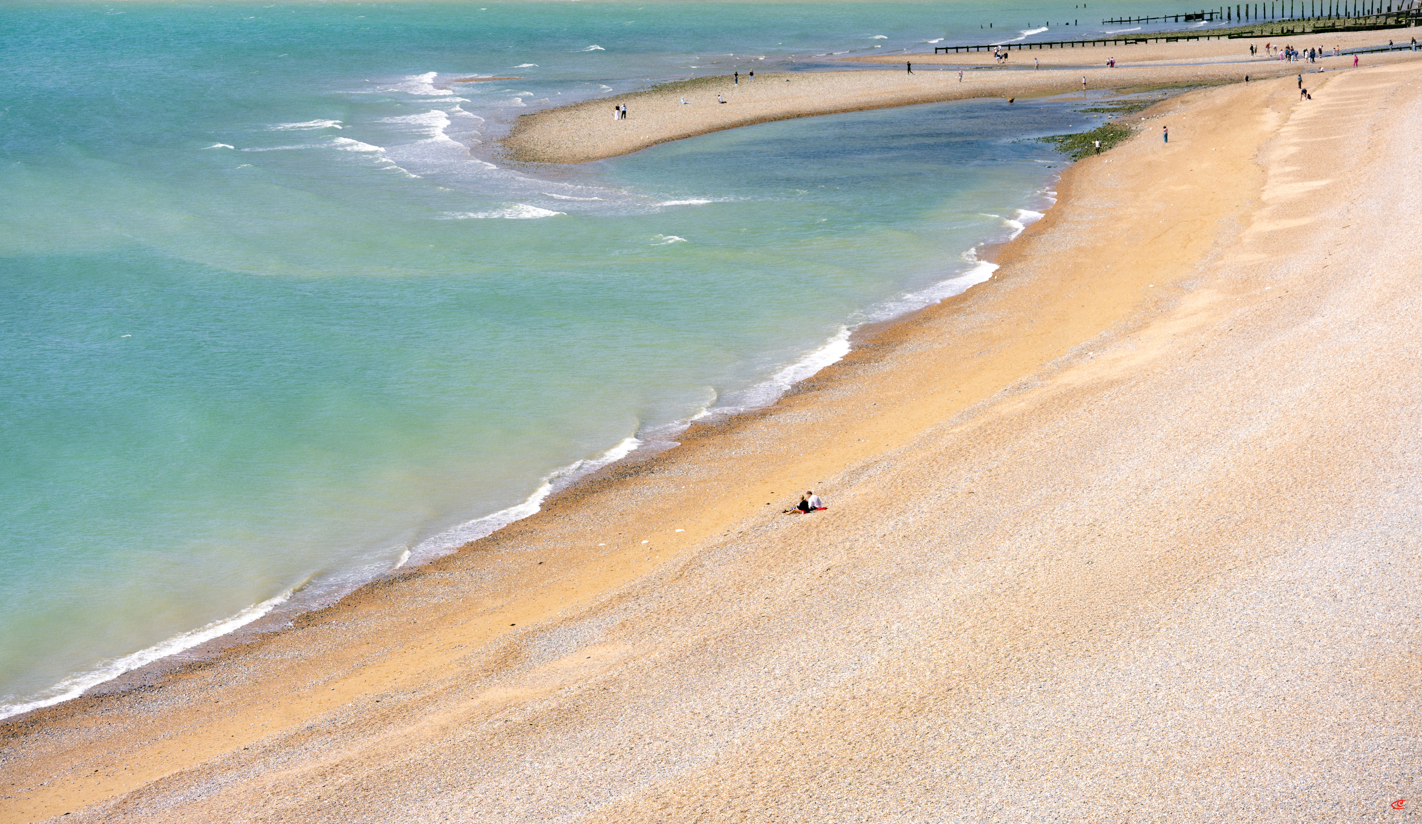 Curving sandy shoreline with pale turquoise water and small waves, a narrow sandbar extending into the sea, and scattered people walking along the beach near a distant pier.