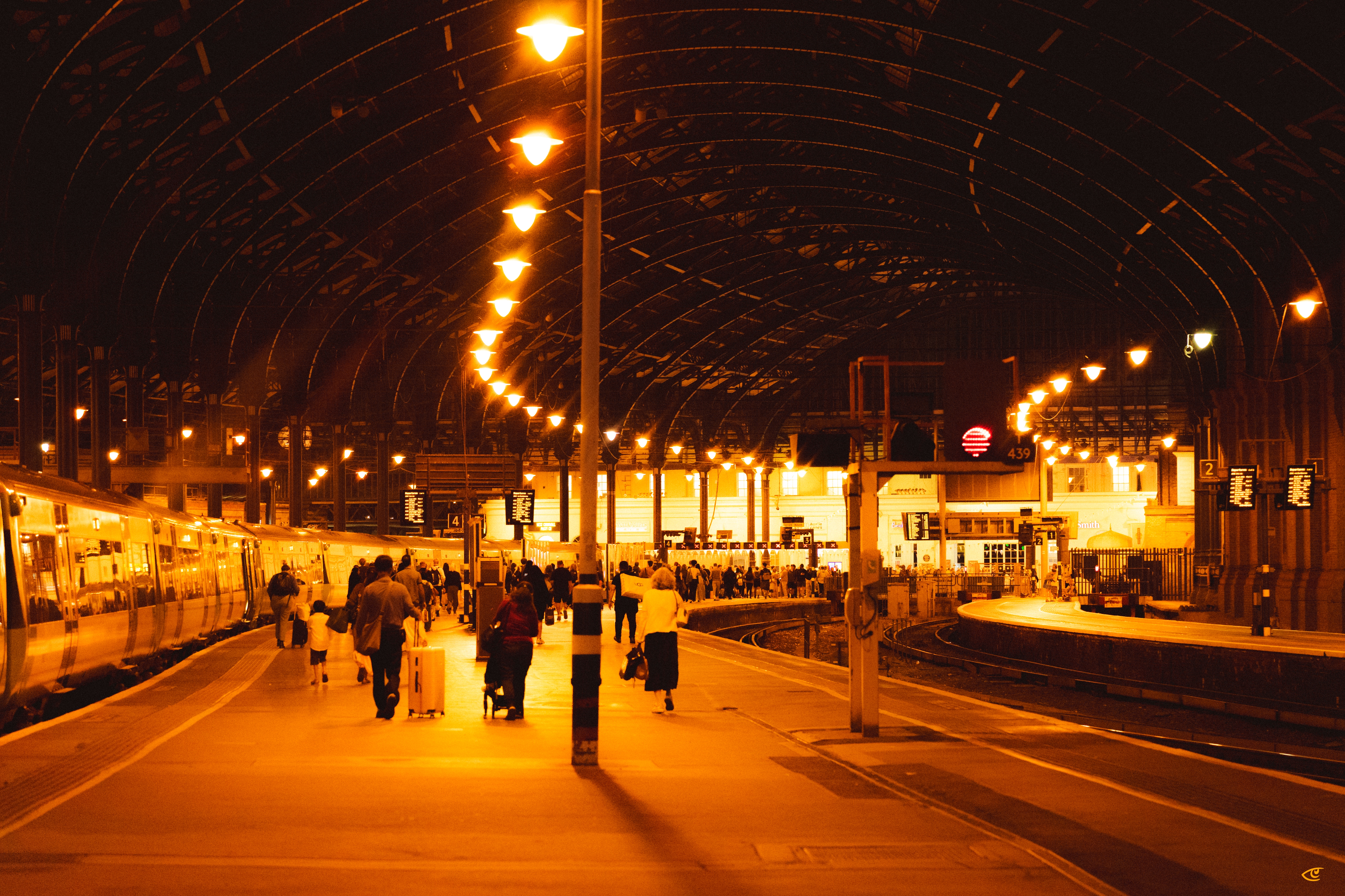 Nighttime view inside a covered train station with an arched roof, rows of warm overhead lights, a train along the left platform, travelers walking with suitcases in the center, and empty tracks curving to the right with platform signs in the background.
